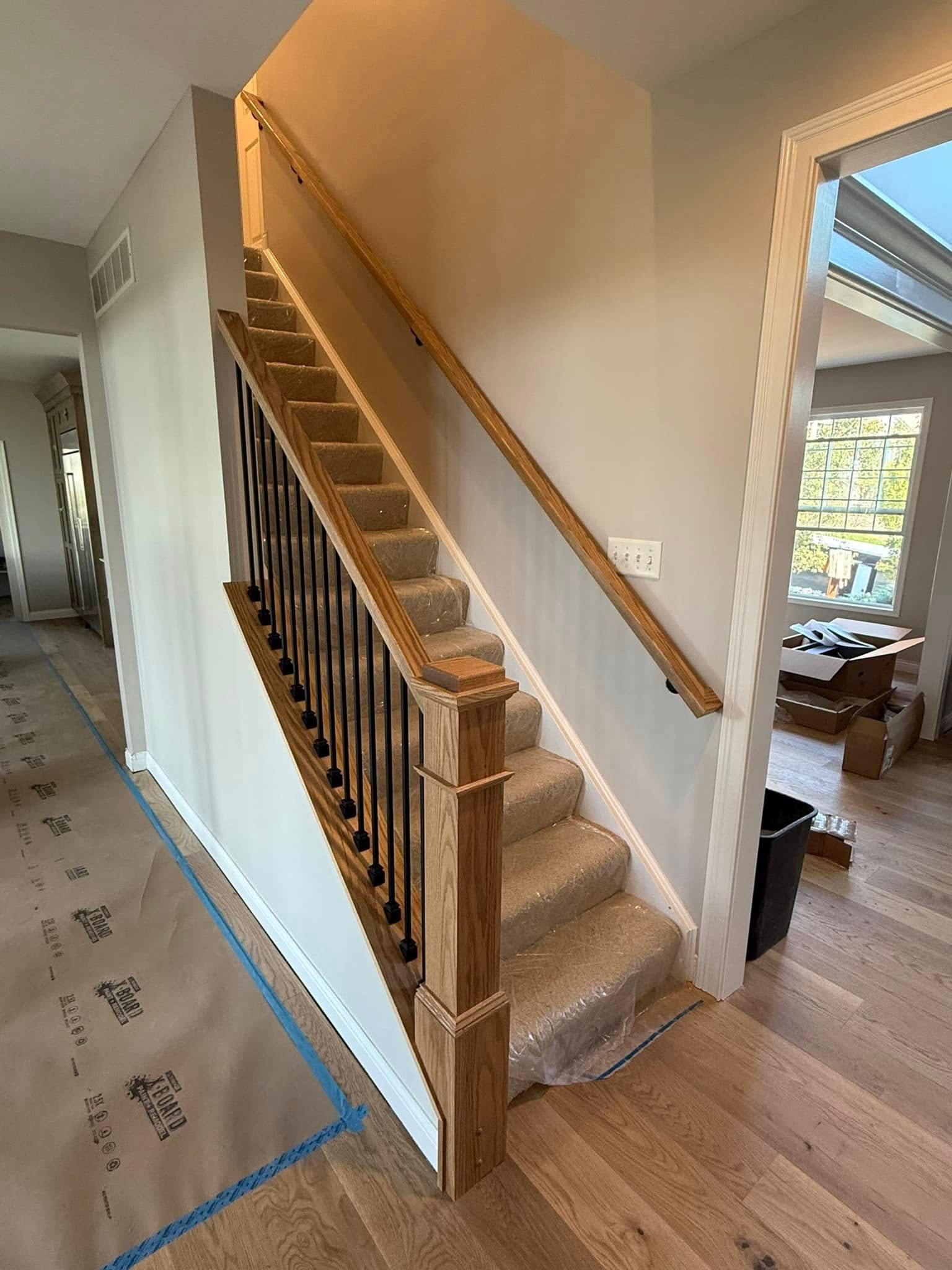 Staircase with wooden handrails and black iron balusters, tan carpeted stairs, and light-colored walls.