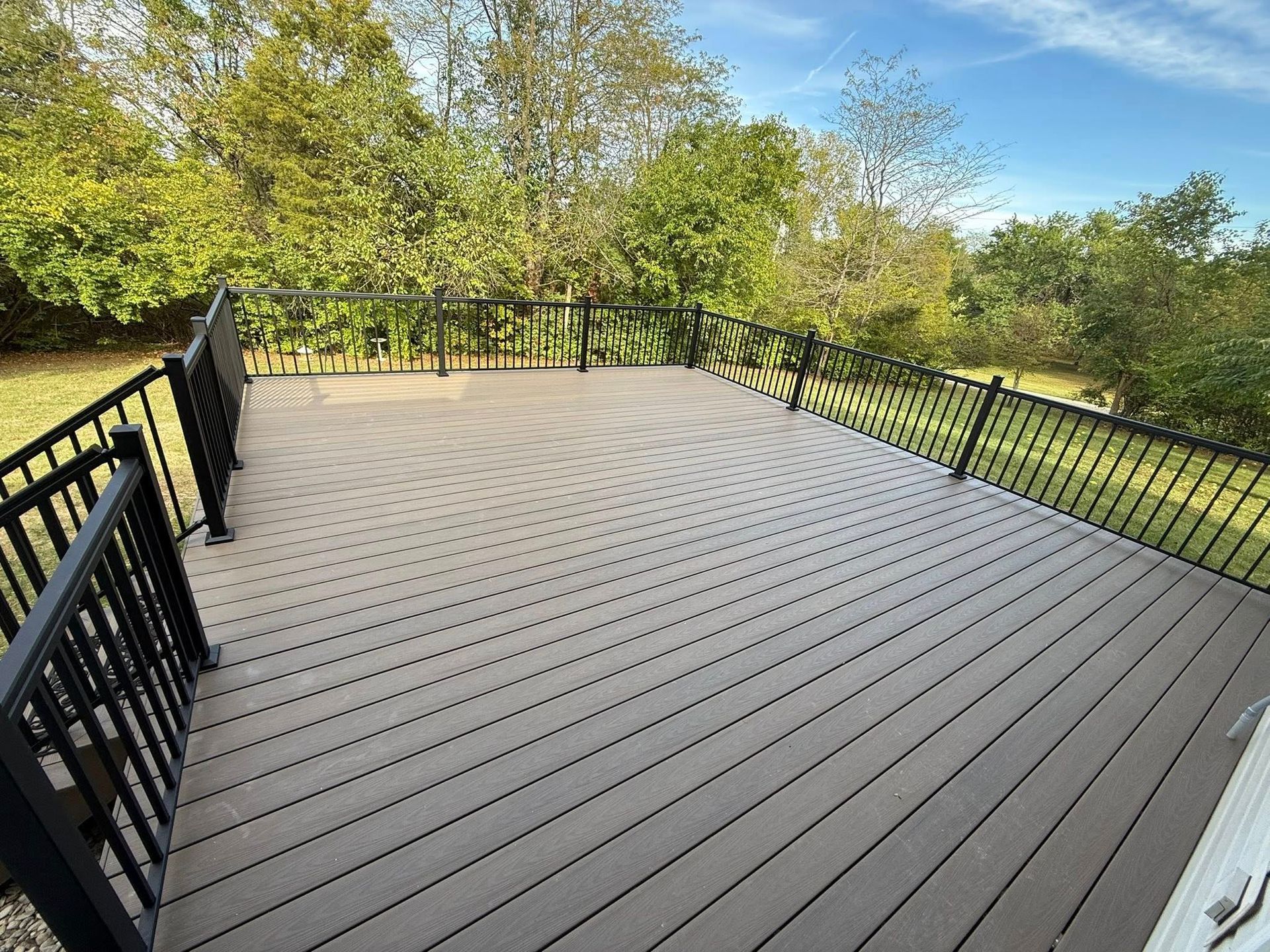 Elevated wooden deck with black railings overlooking a grassy yard and trees under a blue sky.