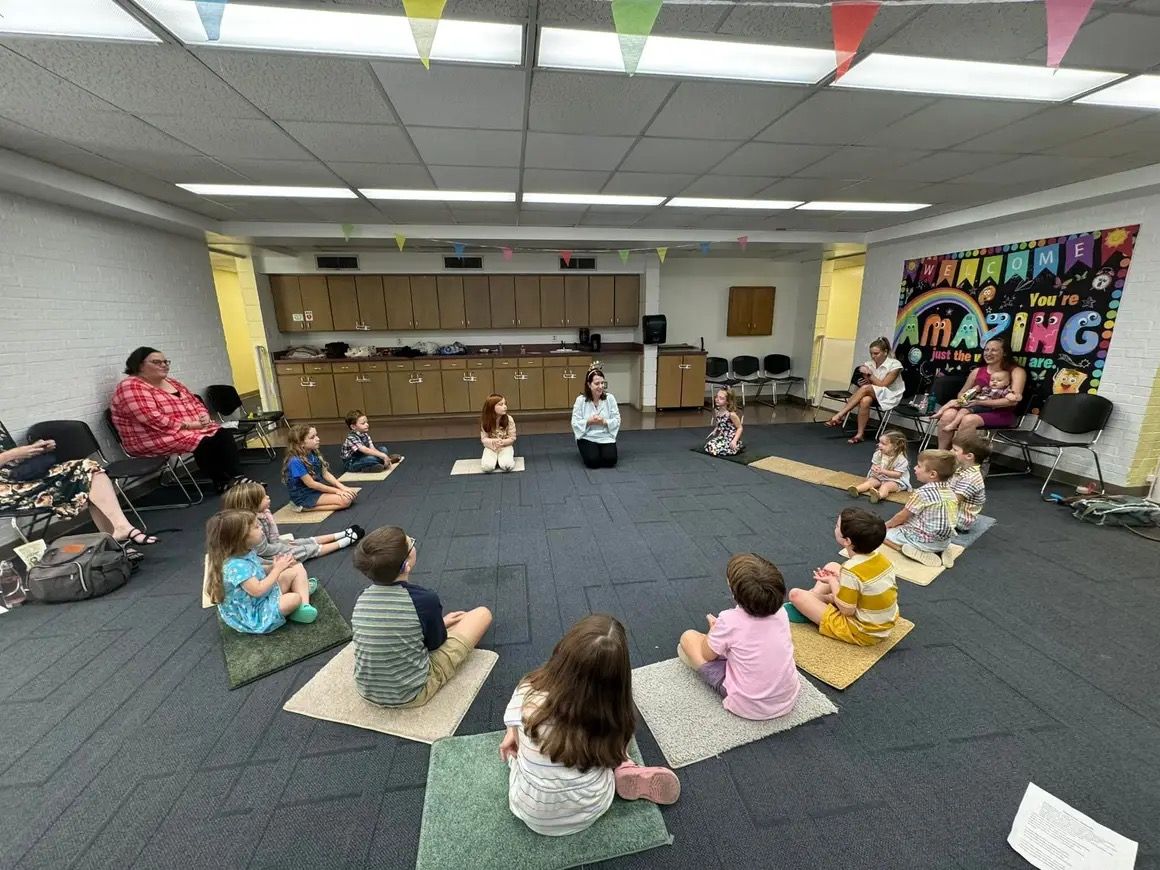 Children seated in a circle on mats in a classroom, facing a teacher in front of a kitchen play area.