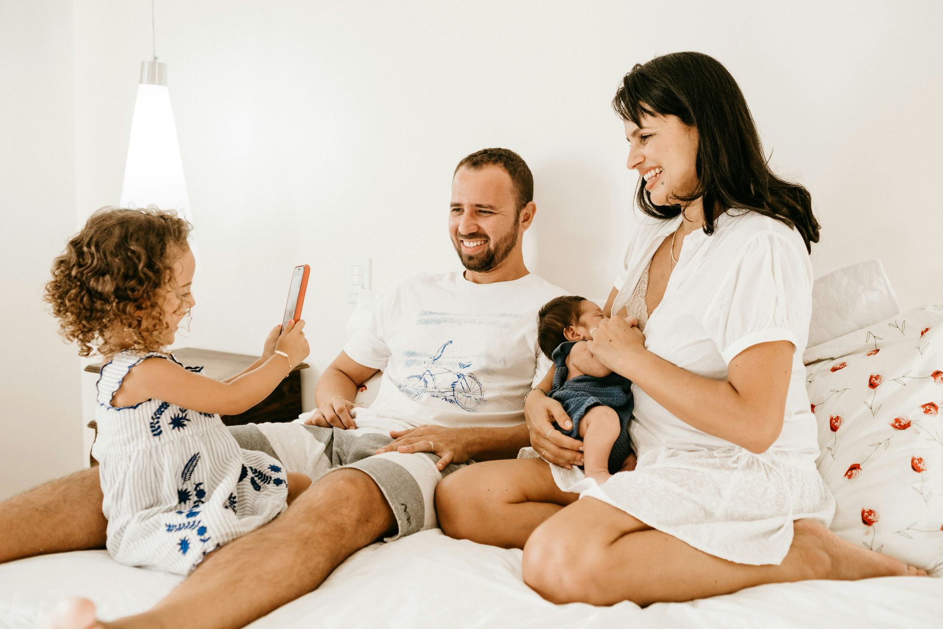 Family on a bed: mother breastfeeding baby, father watches, child holds phone, all smiling in bedroom.