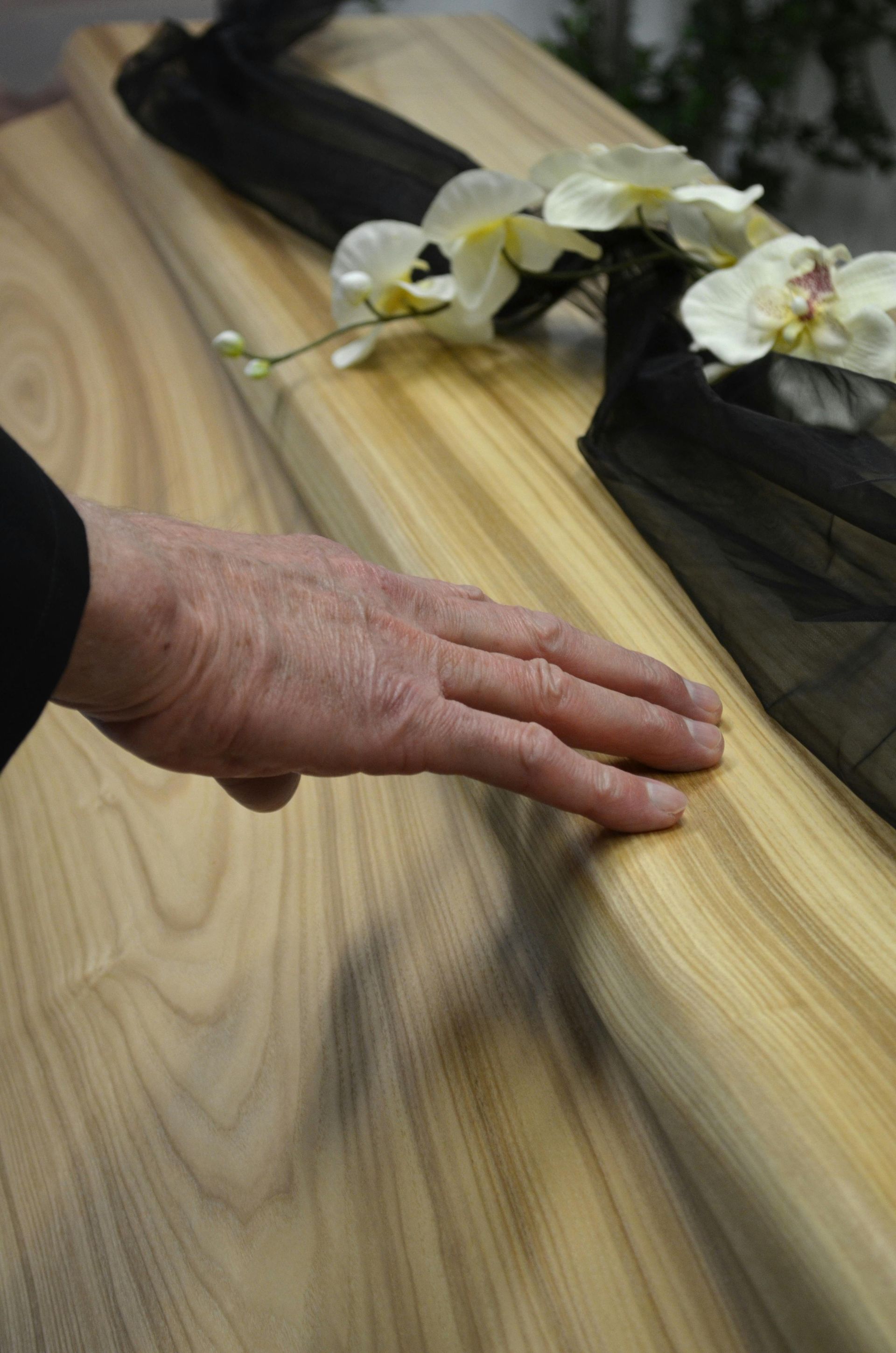 Hand resting on a wooden casket draped with a black cloth and white orchids.