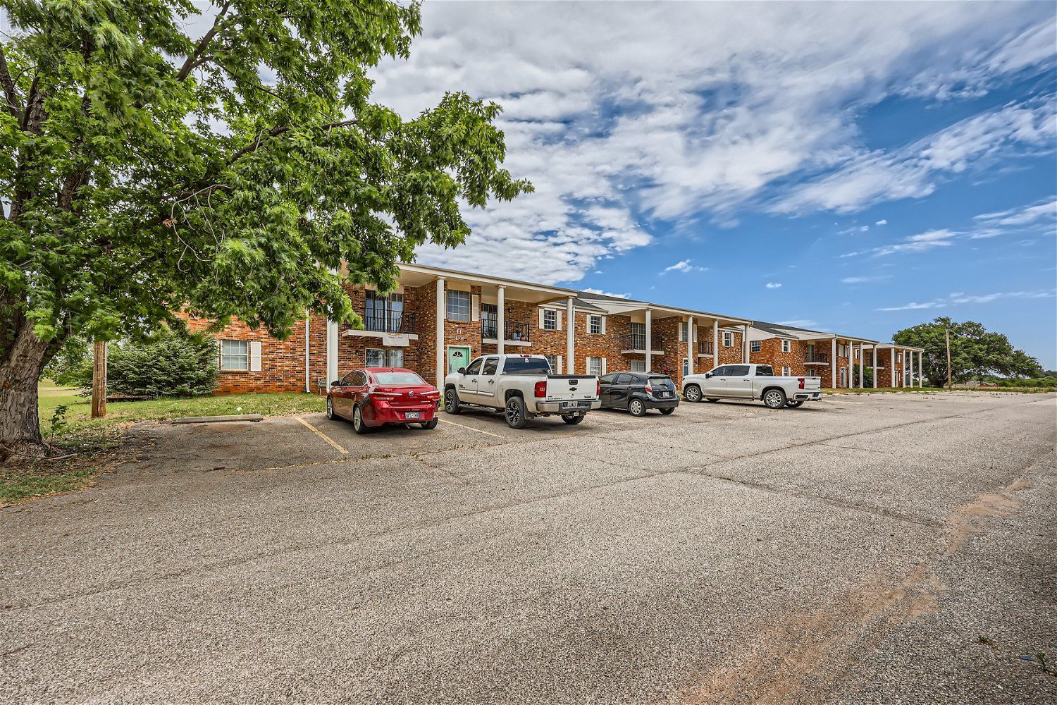 Apartment building with cars parked out front on a gravel lot. Blue sky and trees in the background.