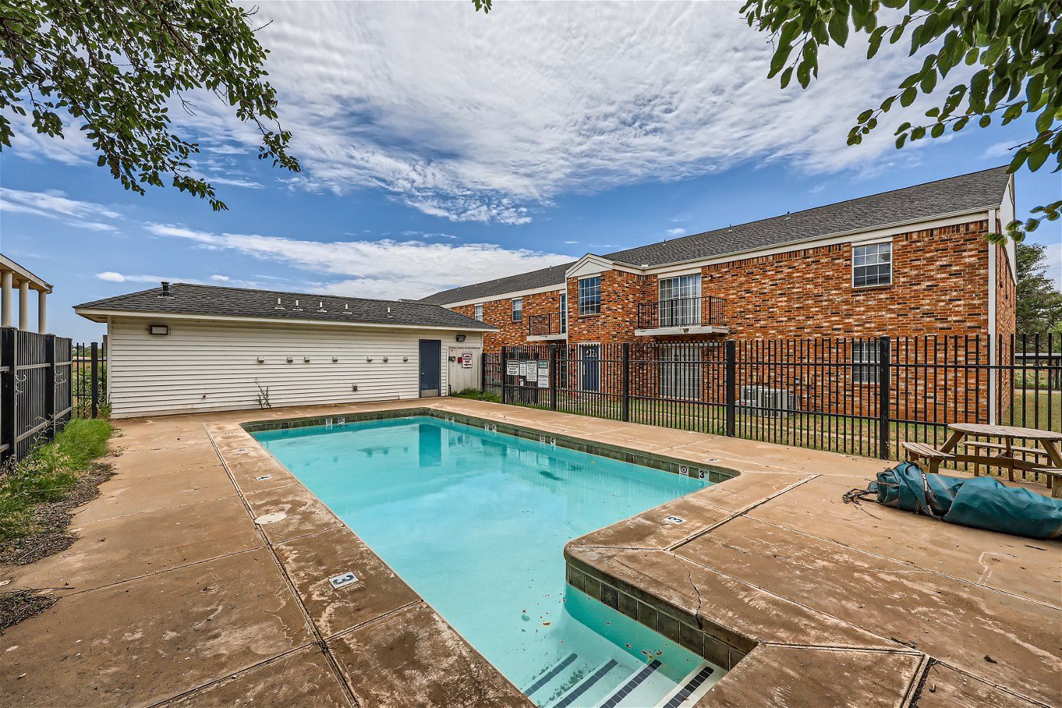 A rectangular swimming pool in front of a brick building. Blue water, concrete decking, sunny day.