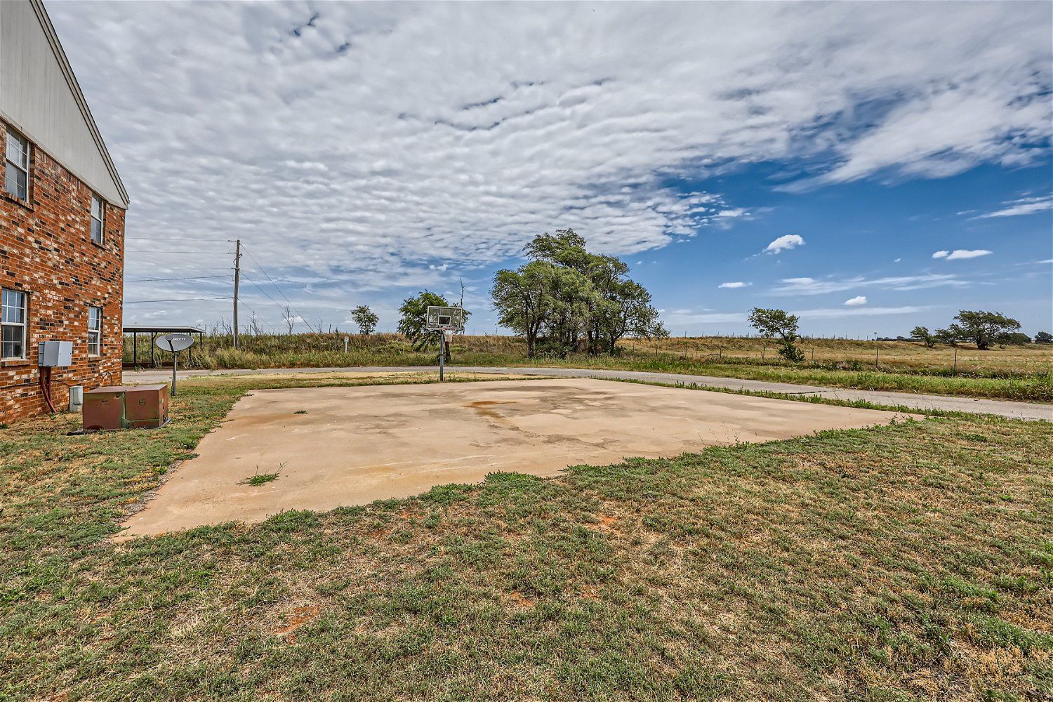 Concrete basketball court with a hoop, surrounded by grass and a brick building, under a cloudy sky.