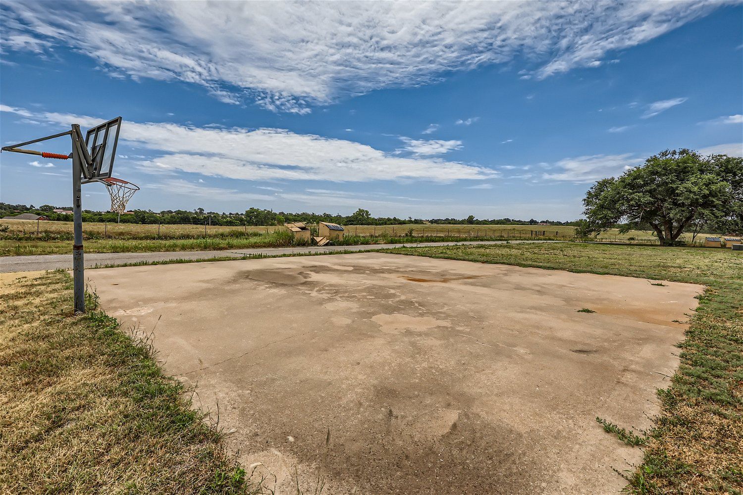 Concrete basketball court, hoop, and backboard set against a field and blue sky.