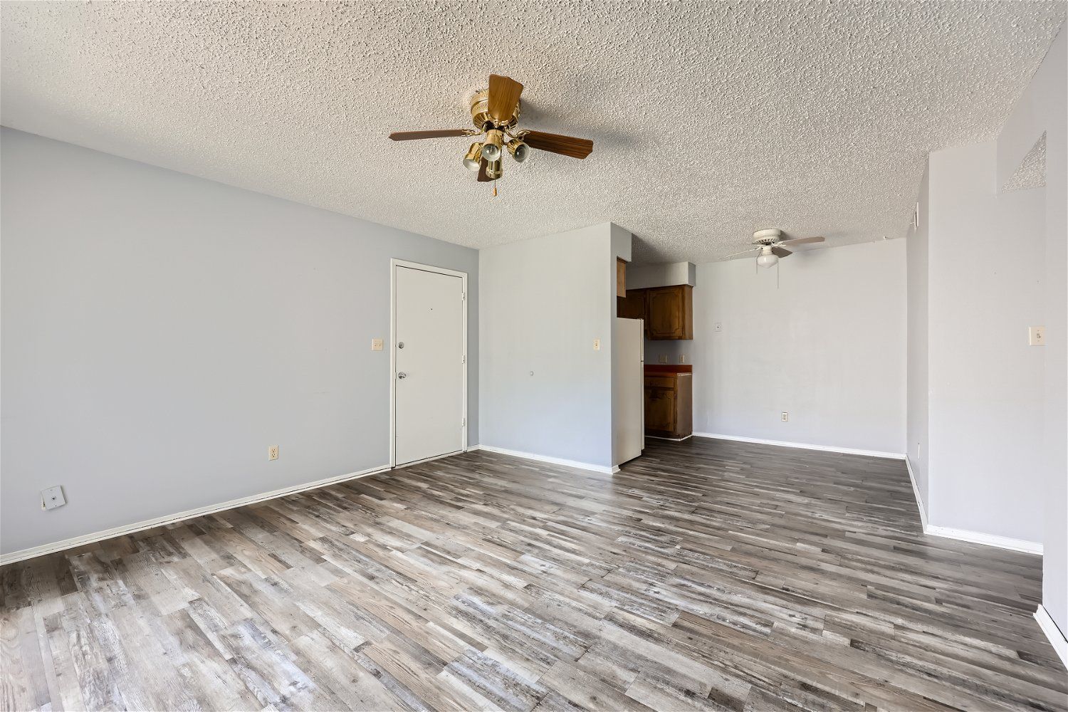 Empty living room with light gray walls, wood-look flooring, popcorn ceiling, and two ceiling fans.