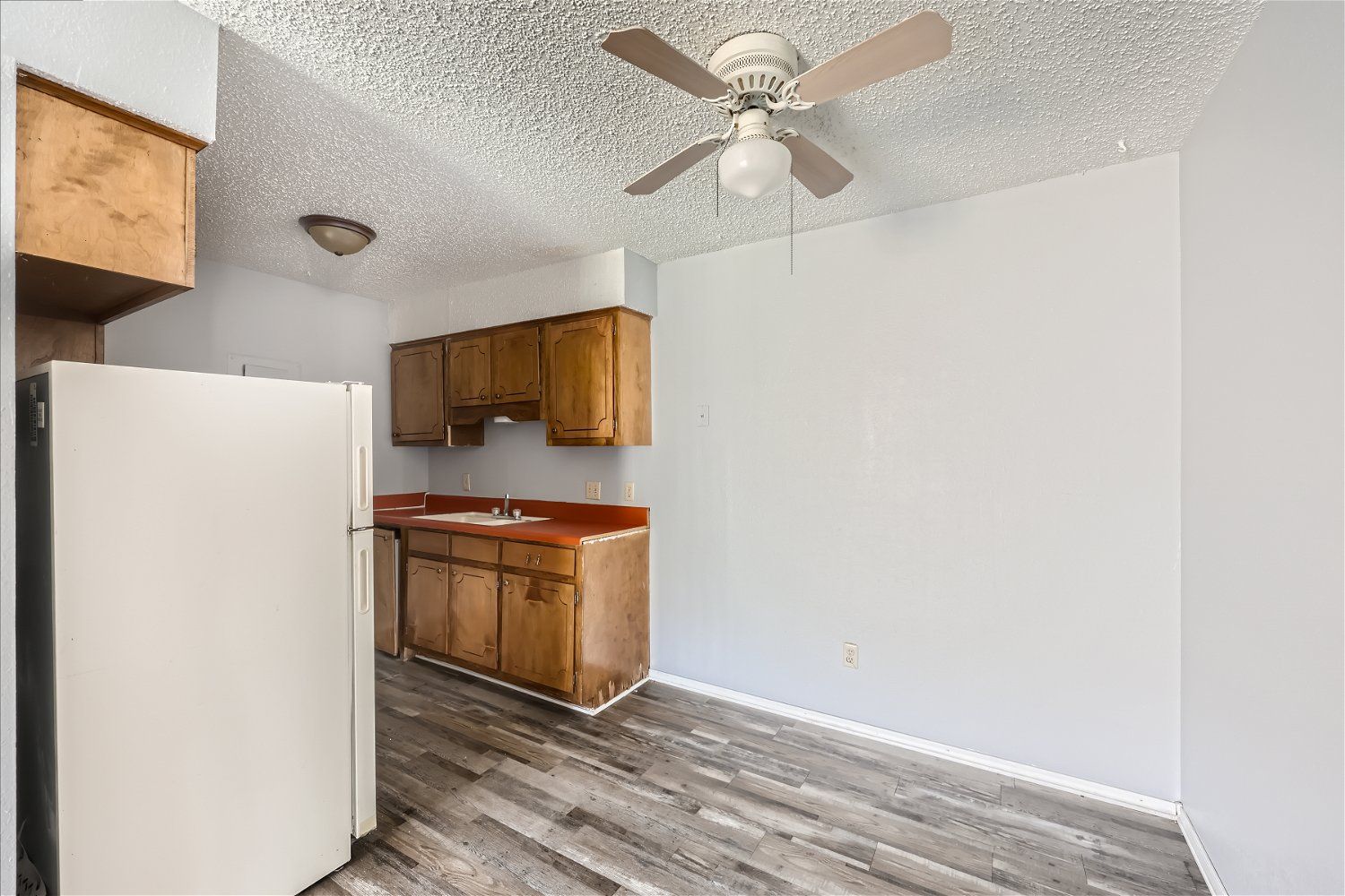 Kitchen with wooden cabinets, white fridge, red countertop, and ceiling fan. Gray walls and flooring.