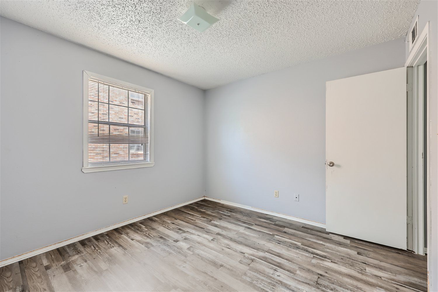 Empty room with light gray walls, window, white door, and wood-look flooring.