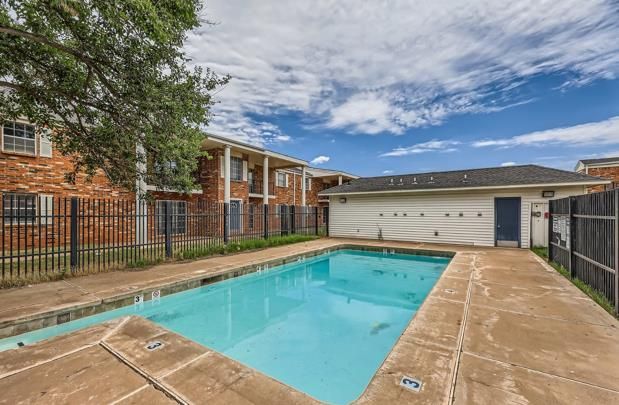 Swimming pool in courtyard of two-story brick apartment buildings, under a partly cloudy sky.