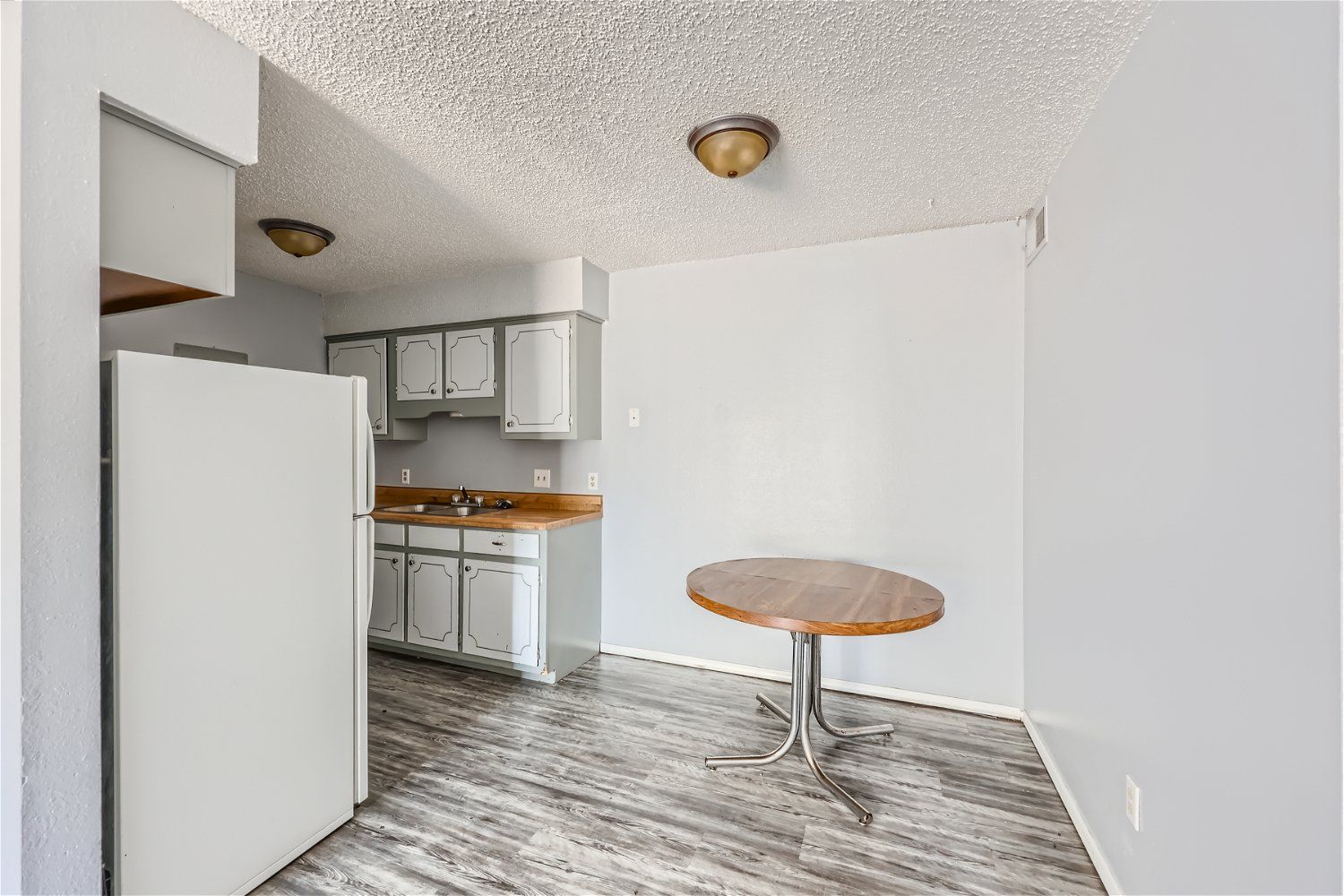 Kitchen with white refrigerator, cabinets, and a round wooden table. Gray flooring, light-colored walls.
