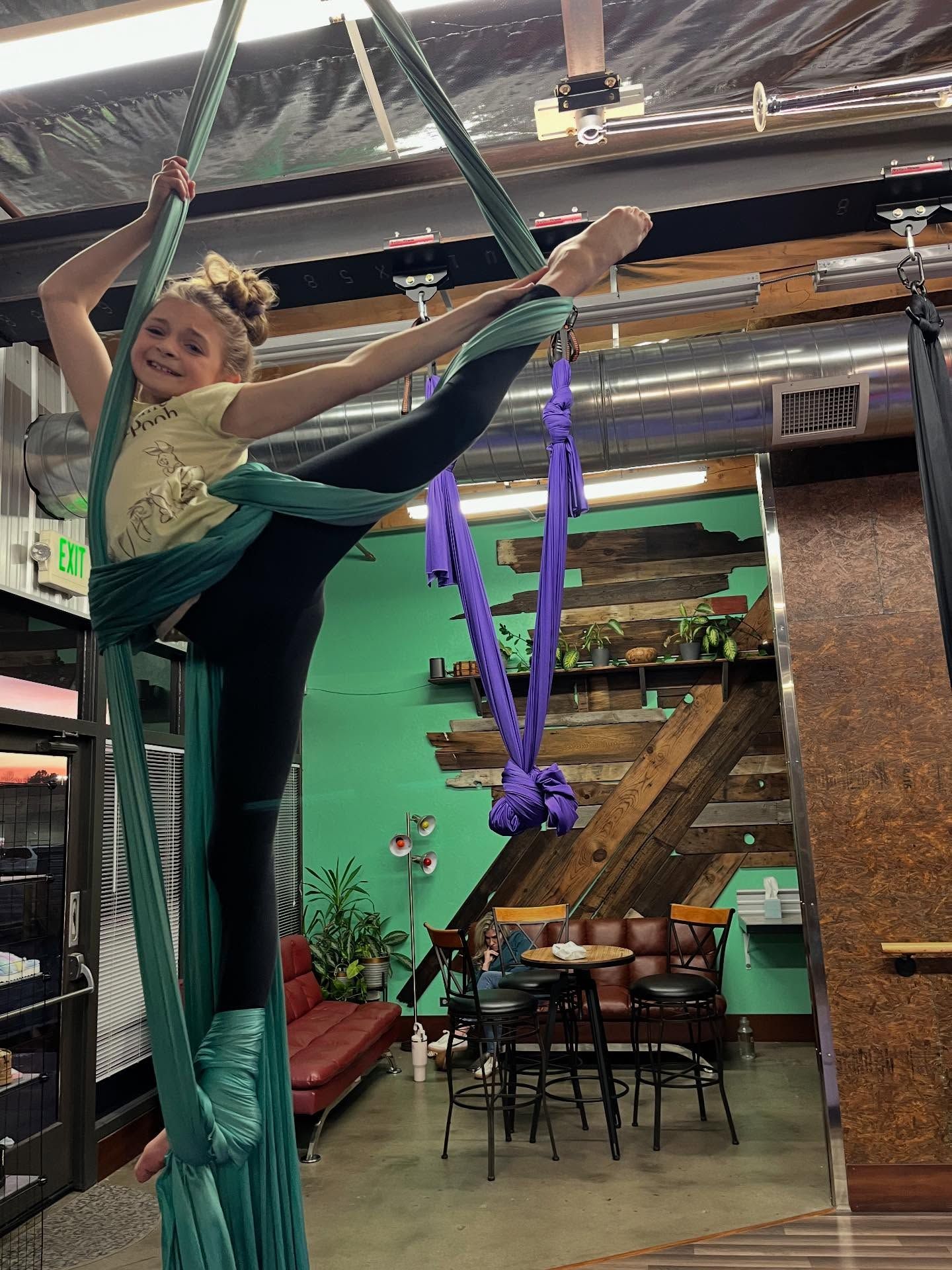 Child in teal aerial silks, posing high indoors at a circus-style studio with green walls and wooden stairs.