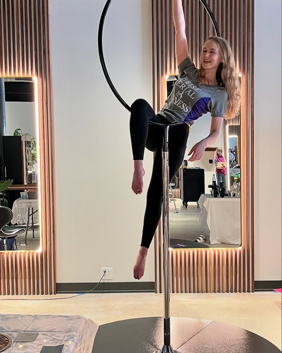 Person performing an aerial hoop pose in a mirrored studio, wearing black leggings and a patterned top.