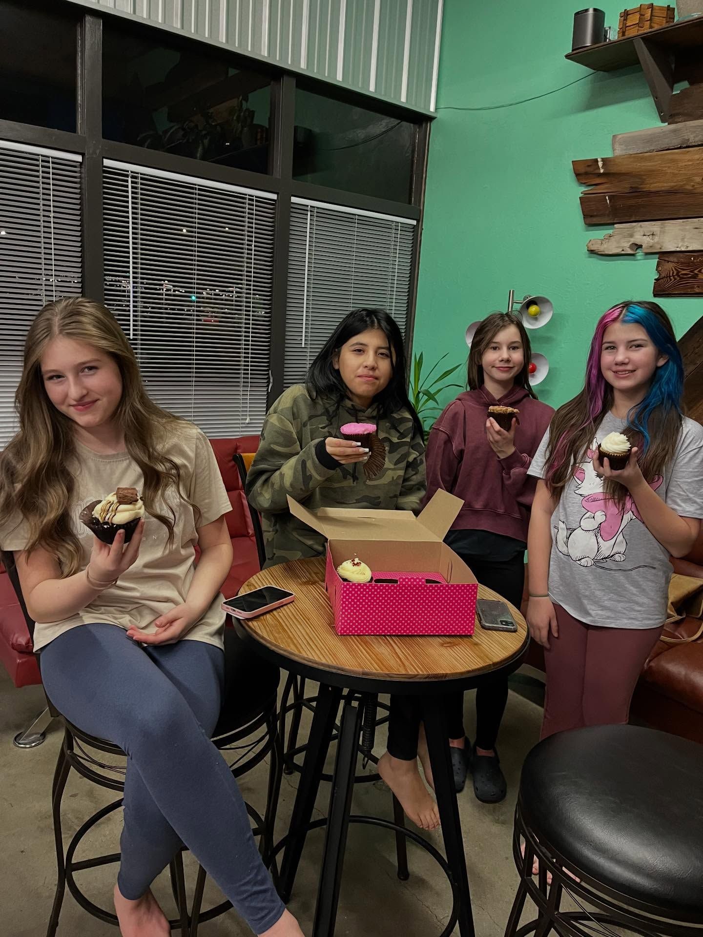 Four people holding cupcakes in a colorful bakery, standing and sitting around a small table