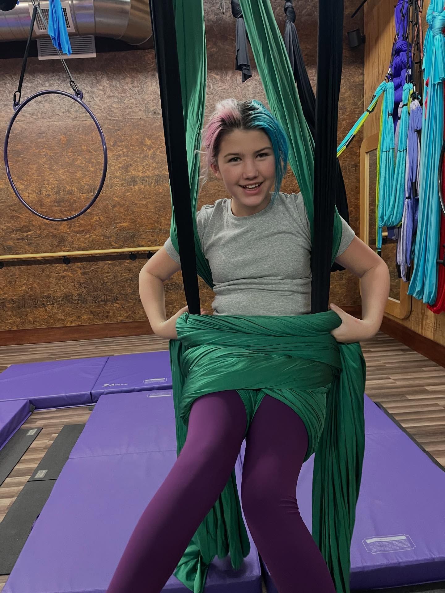 Person seated in green aerial silks on a purple mat, wearing a gray top and purple leggings.