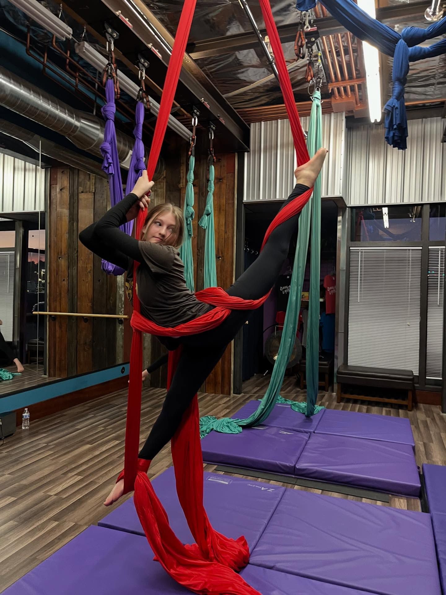 Person performing an aerial silk pose in a studio with red and blue silks and purple mats