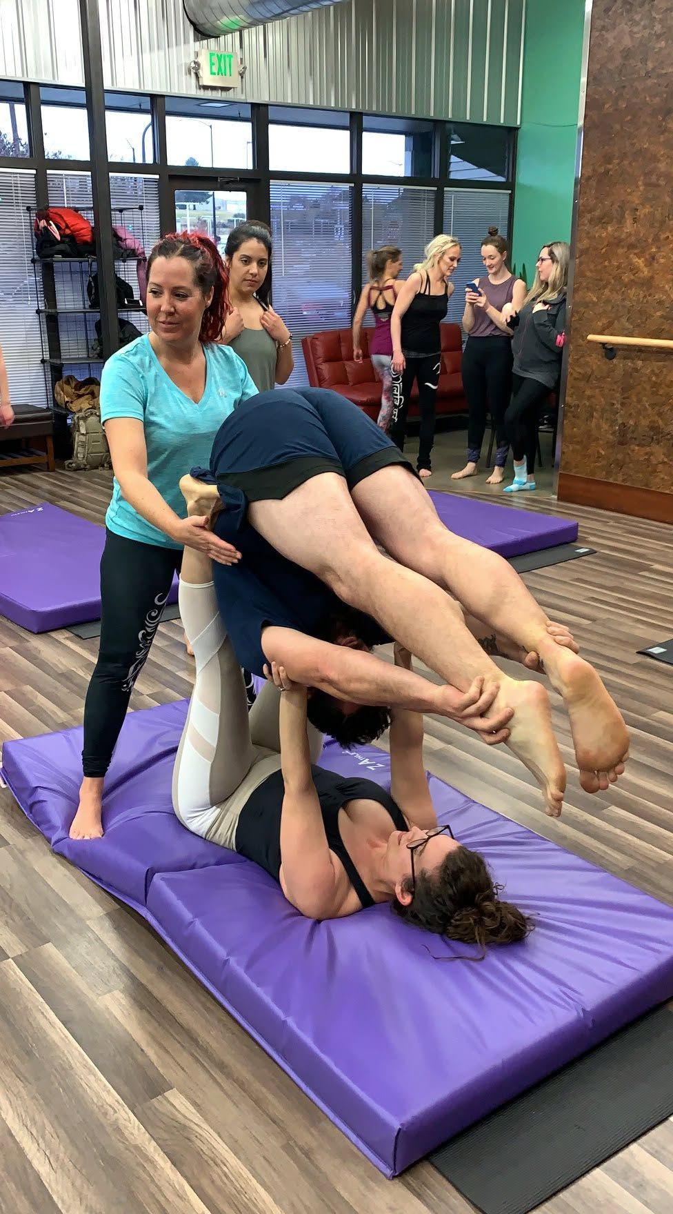 Three people practicing acro yoga on purple mats in a studio, with others watching in the background.