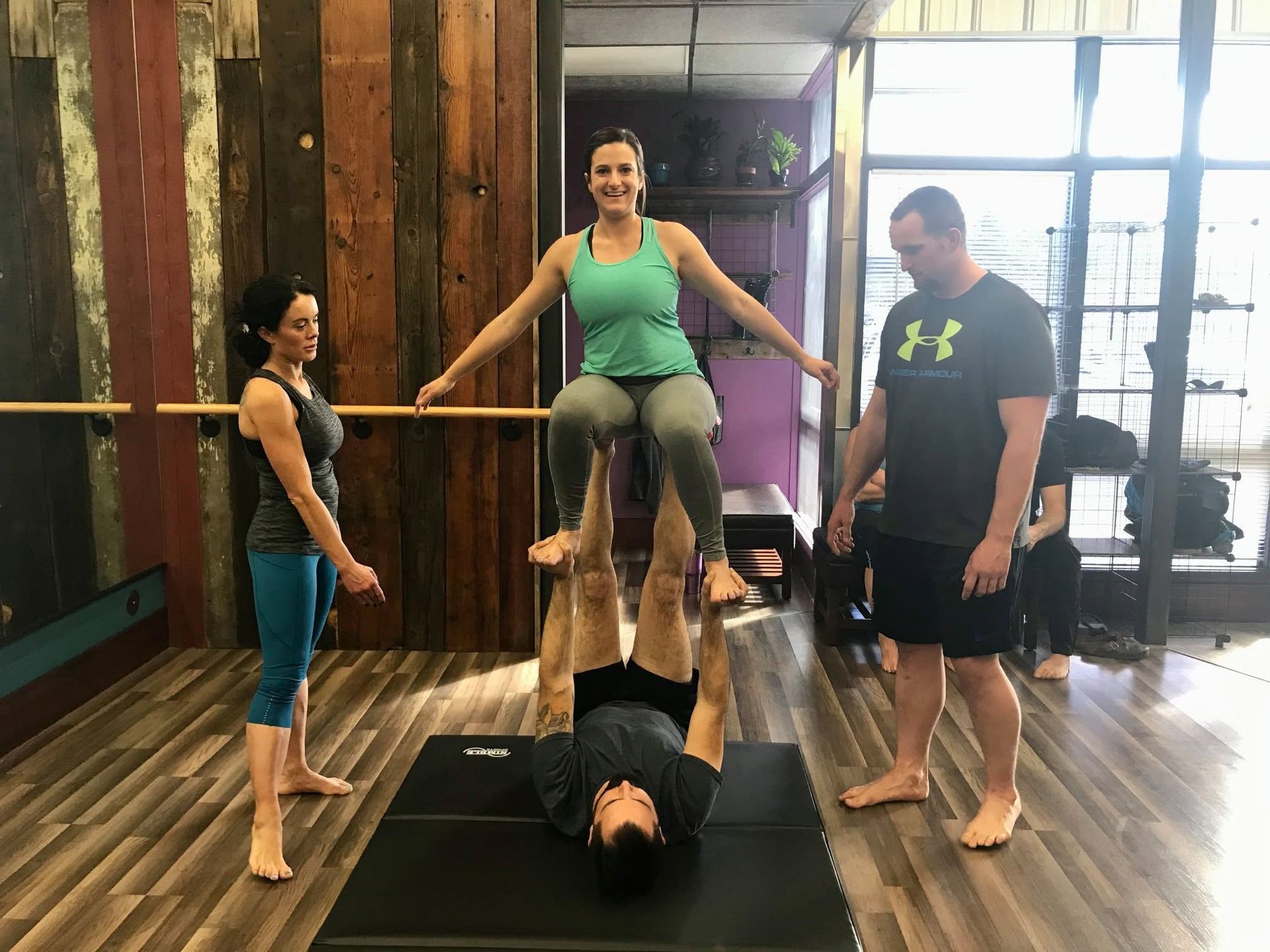 Four people doing a yoga balancing pose in a gym, with one person lying on the floor supporting another.