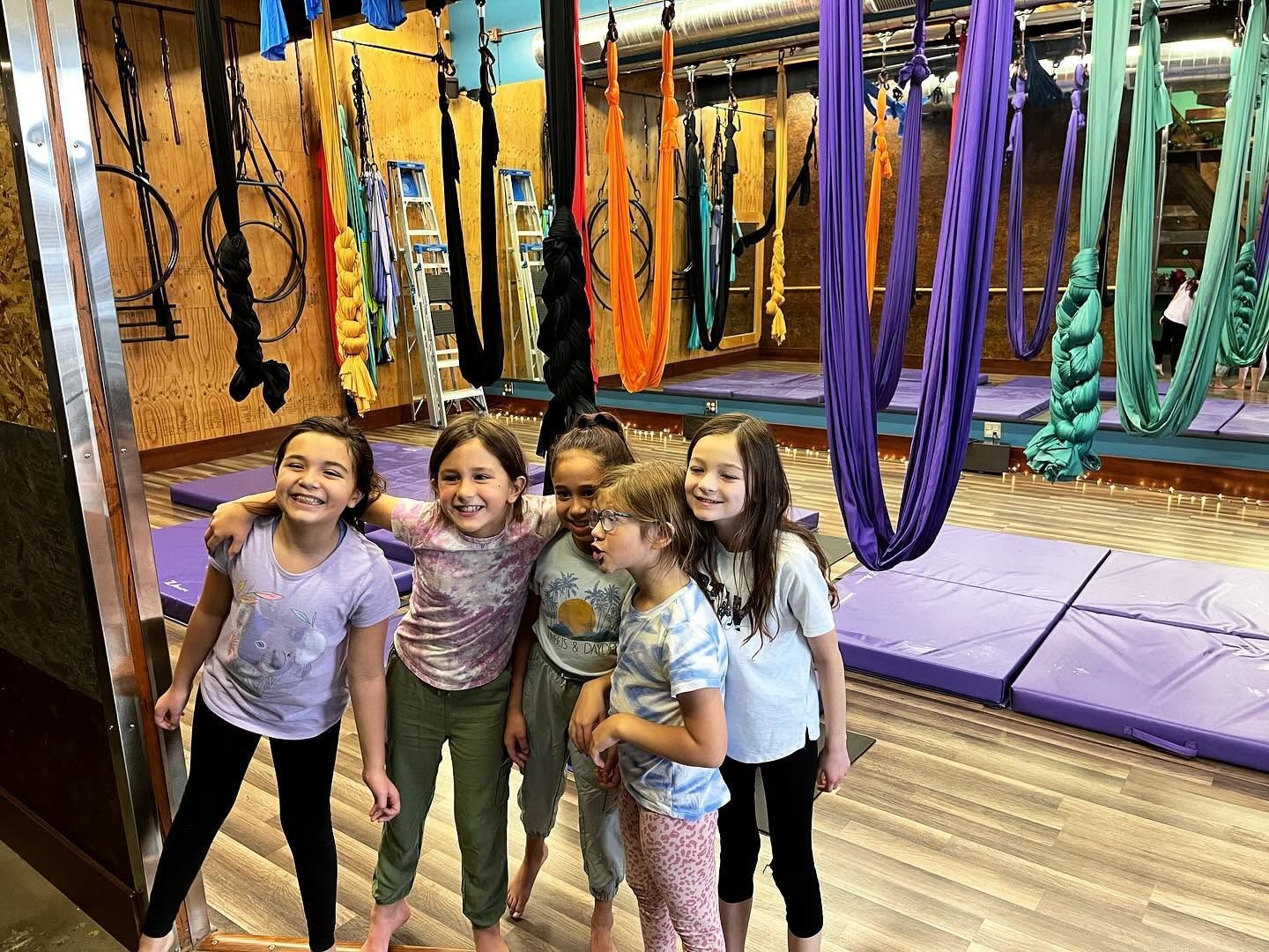 Four smiling kids posing in a colorful indoor gym with hanging ropes and padded floors.