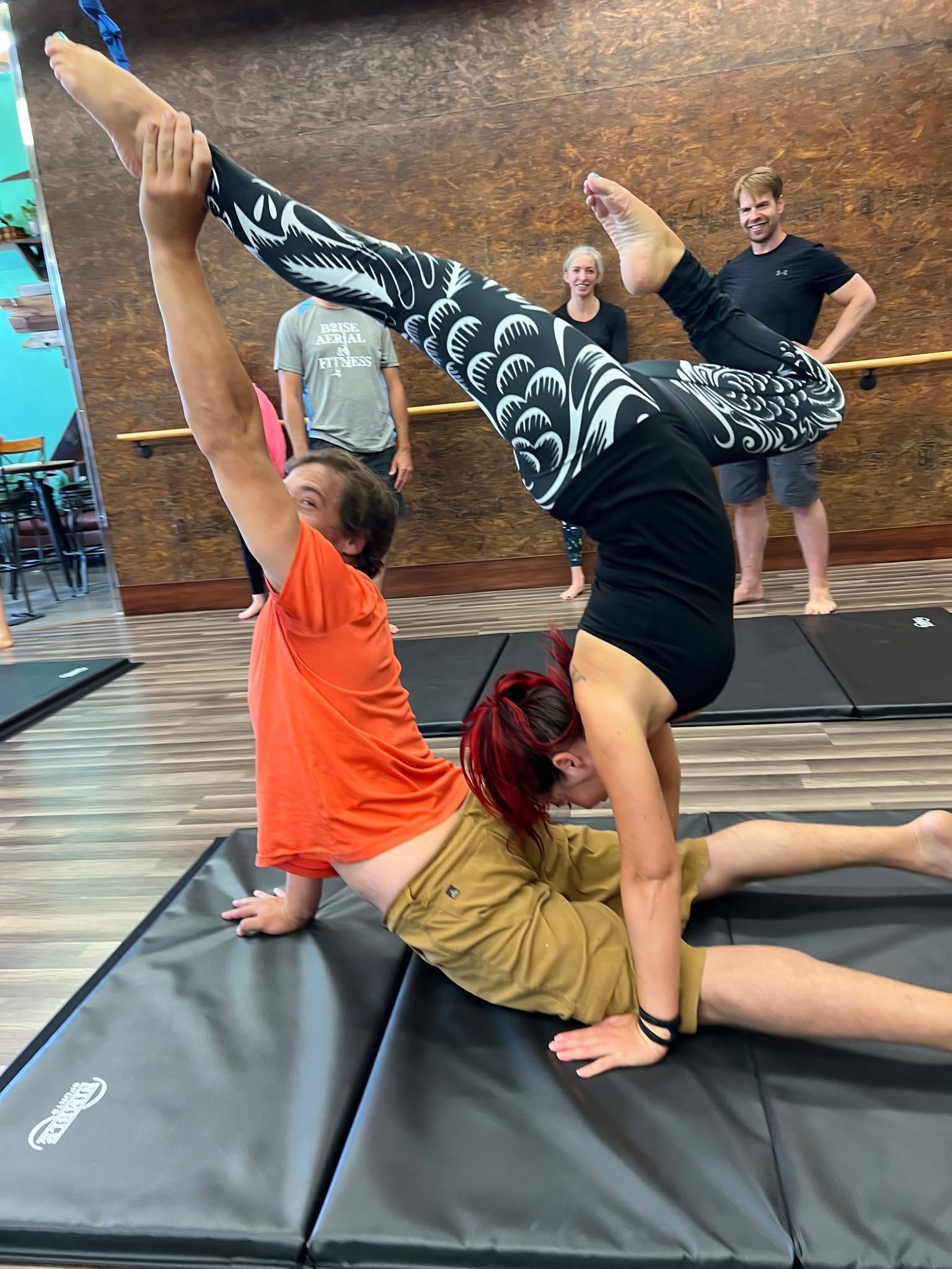 Two people balancing in a yoga pose on mats, with a third person supporting them in an indoor studio.