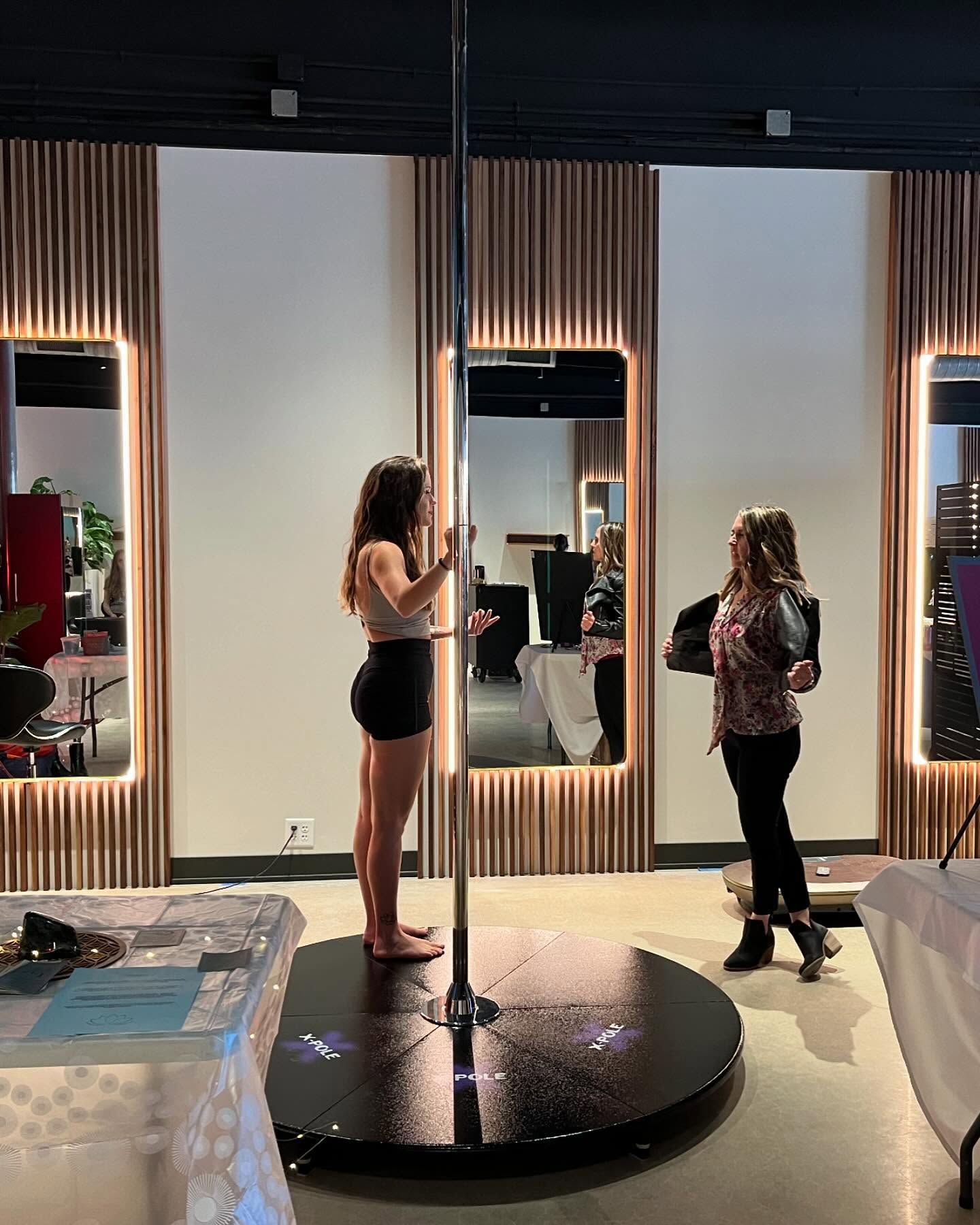 Two women on a pole dance studio stage with mirrors and wood-paneled walls
