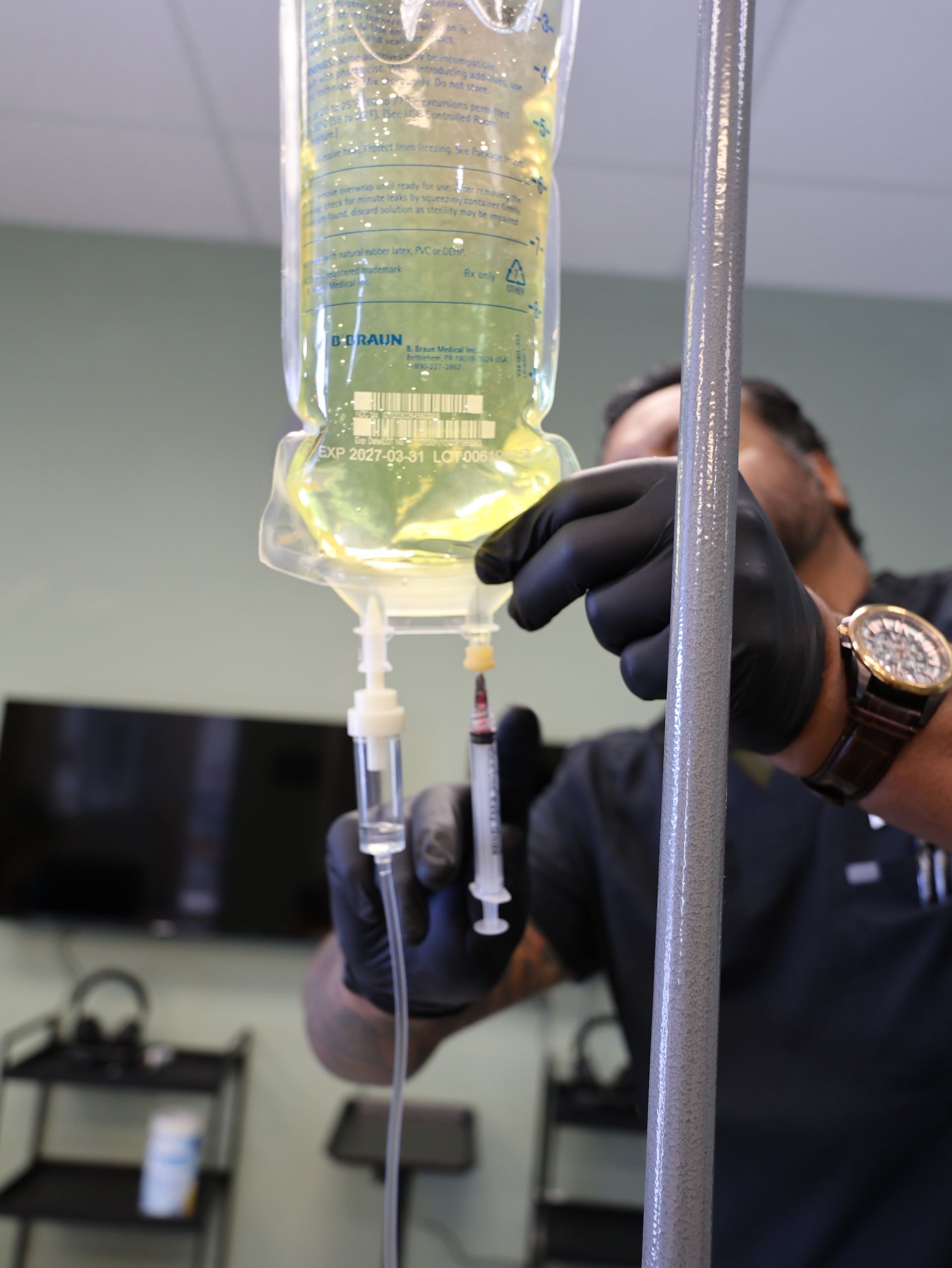 Person in black gloves preparing IV bag with a syringe, in a medical setting. Level Up Falcon