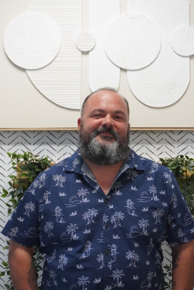 Man with a full beard wearing a blue patterned shirt smiles in front of abstract white art and plants. Level Up Falcon