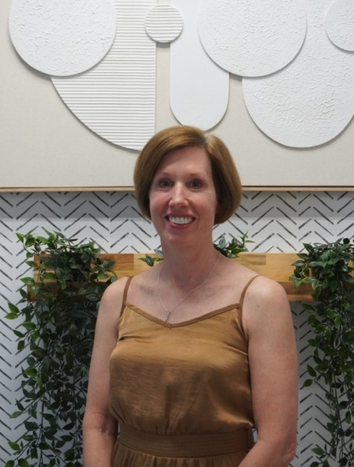 Woman with short hair smiles, wearing a brown top, in front of a wall with abstract art and plants. Level Up Falcon