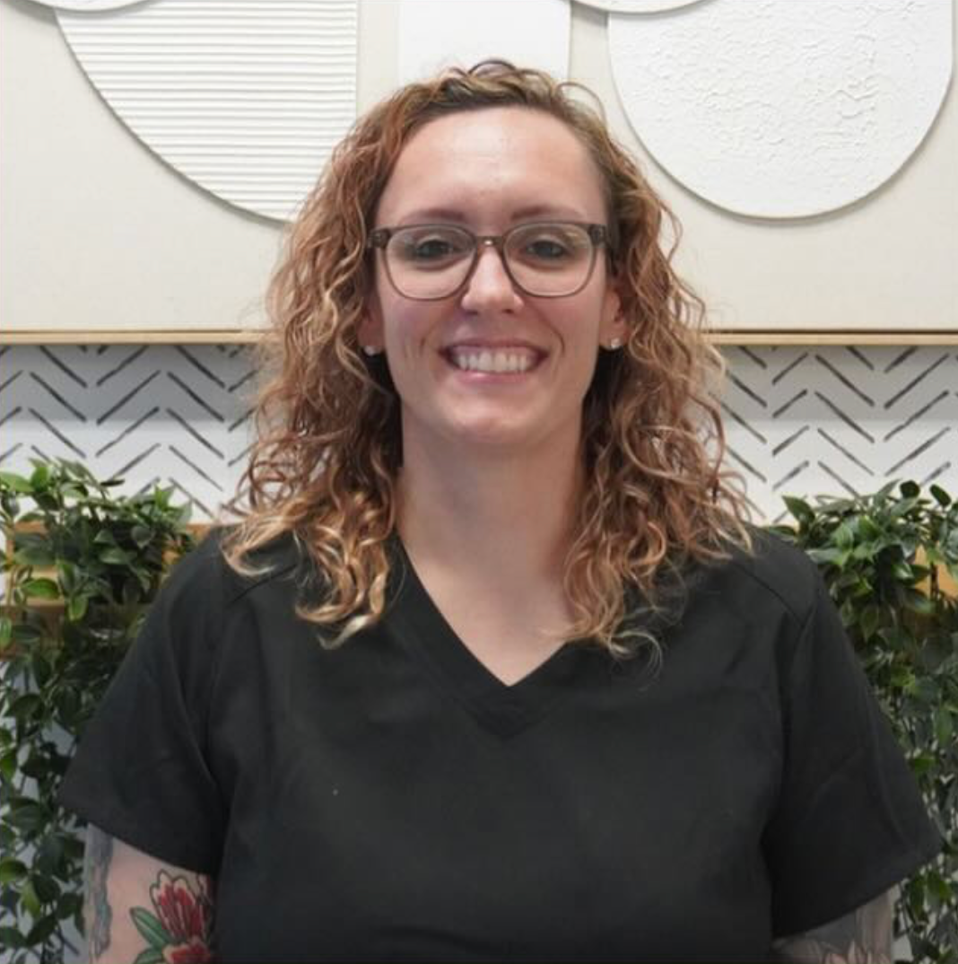 Woman with curly brown hair, glasses, and a black v-neck shirt smiles, surrounded by greenery and art. Level Up Falcon