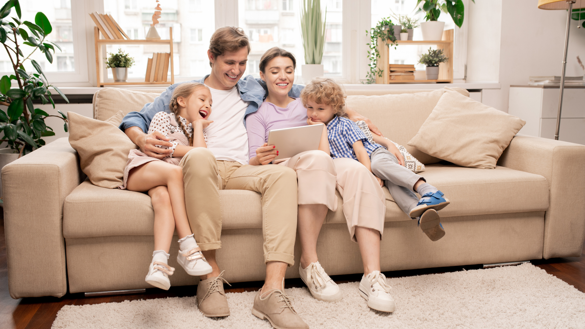 Family of four on a beige sofa, looking at a tablet. Smiling. Living room with plants.