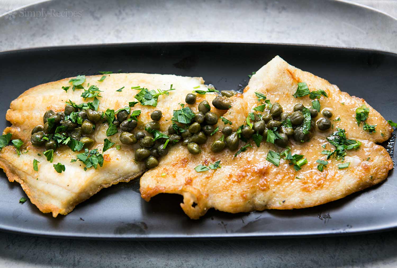 Dos trozos de pescado con alcaparras y perejil en un plato negro.