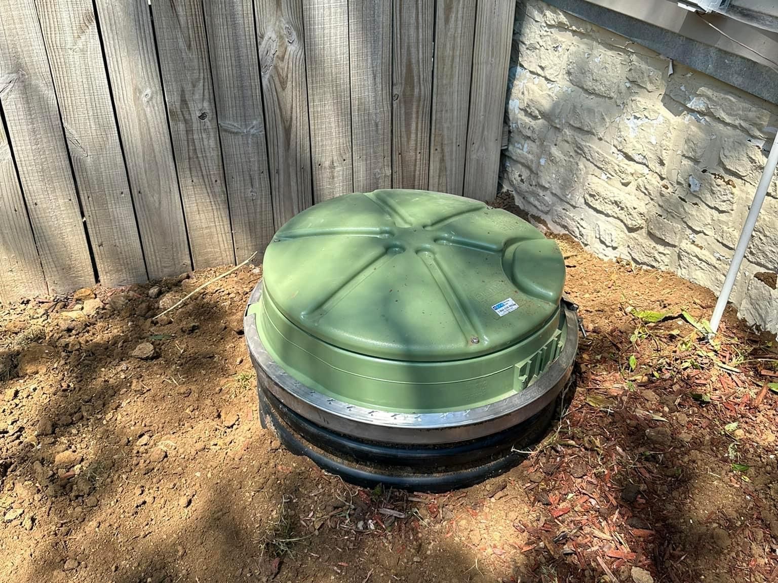 A green drain cover is sitting in the dirt next to a wooden fence.