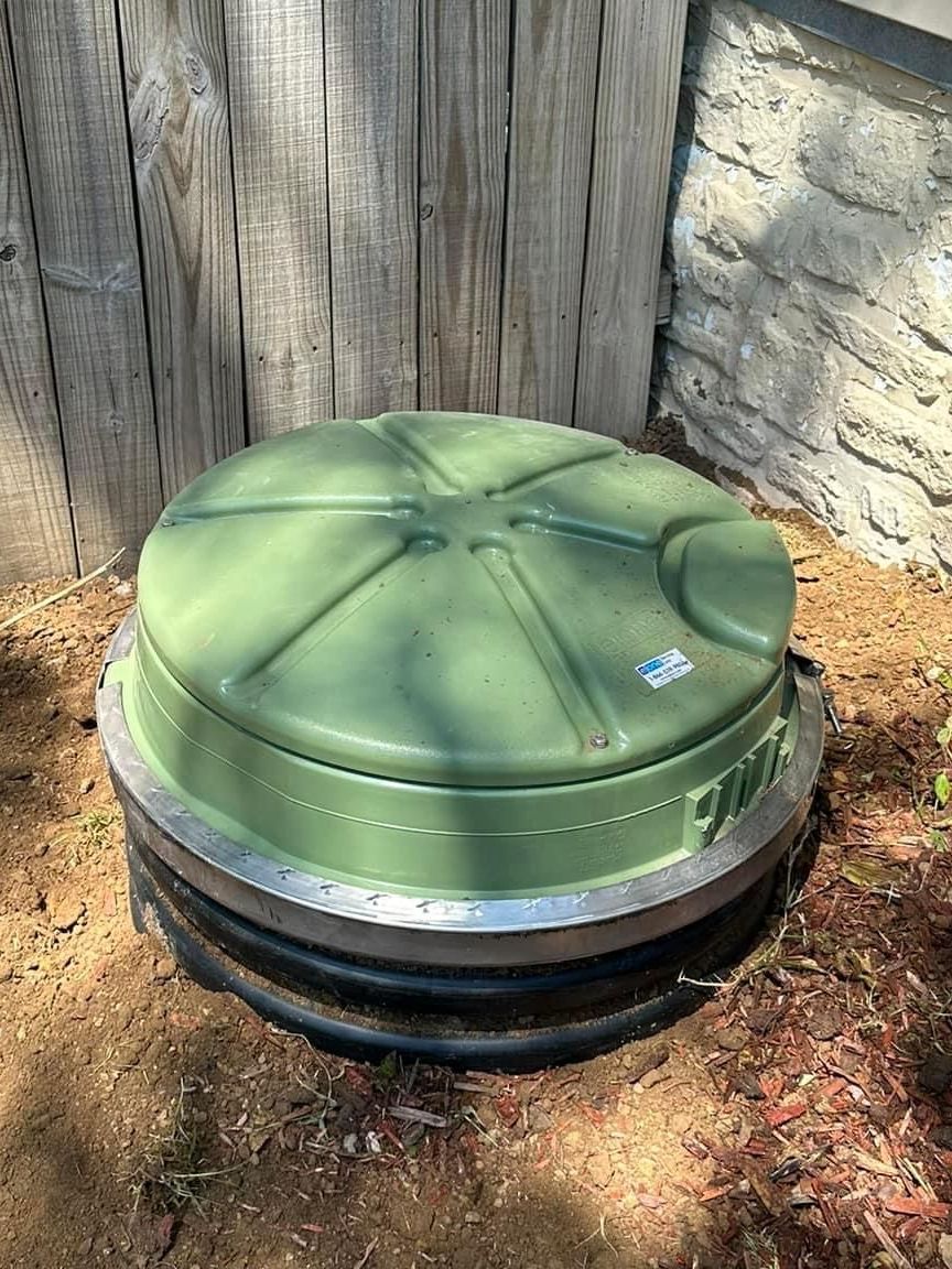 A green drain cover is sitting in the dirt next to a wooden fence.