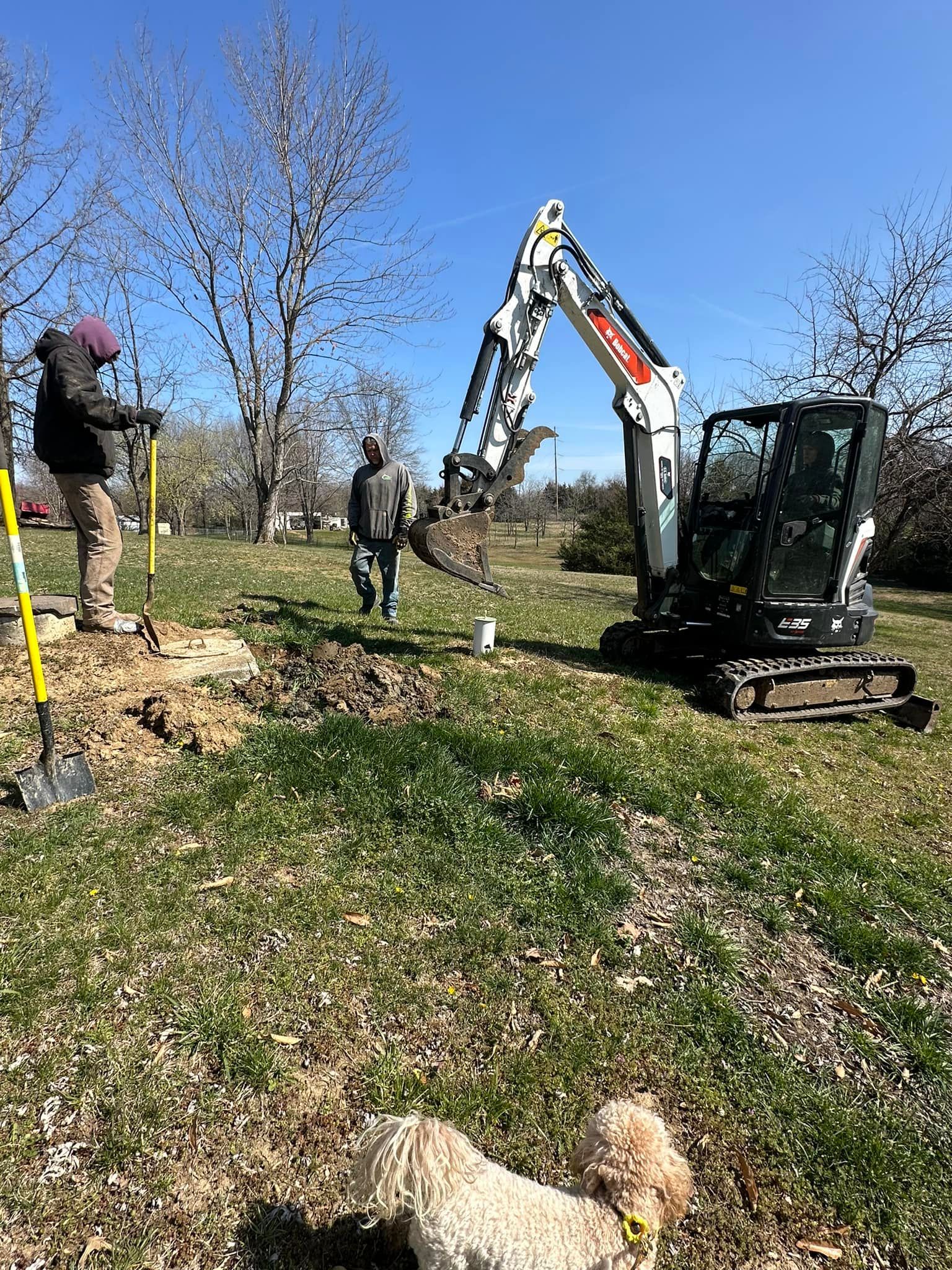 A man and a dog are standing next to an excavator in a field.