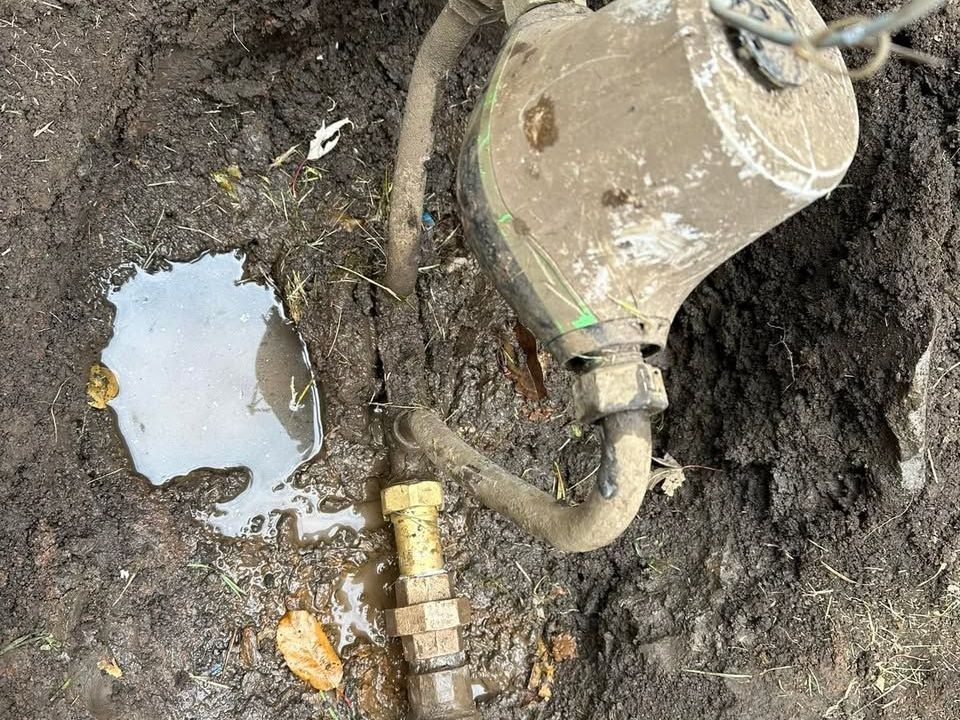 A muddy water meter is sitting in the dirt next to a puddle of water.