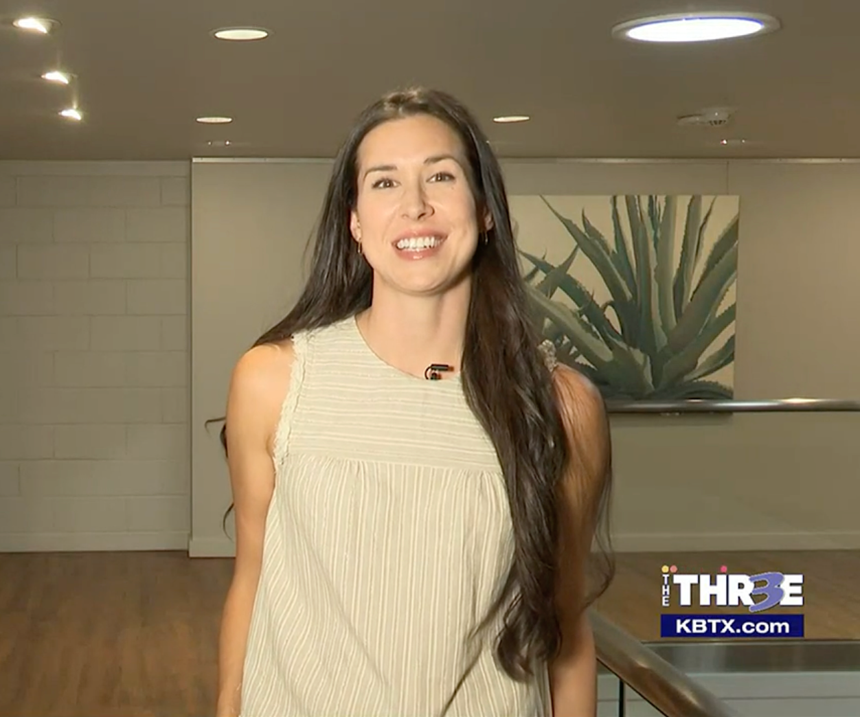 A woman with long hair is smiling in front of a wall with a picture of a plant on it.