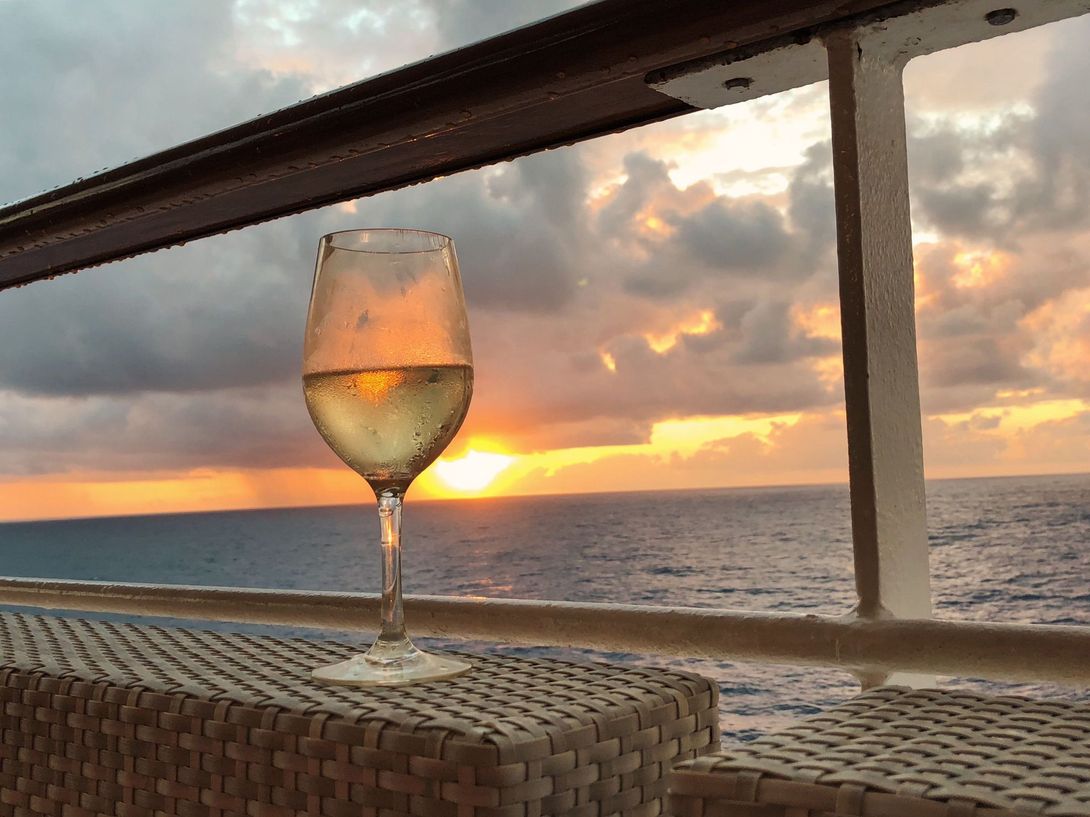 Glass of white wine on a wicker table, ocean sunset in background.