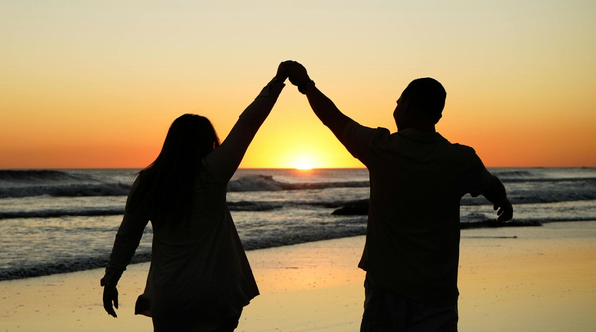 Couple holding hands in a celebratory gesture as they watch a sunset on the beach.
