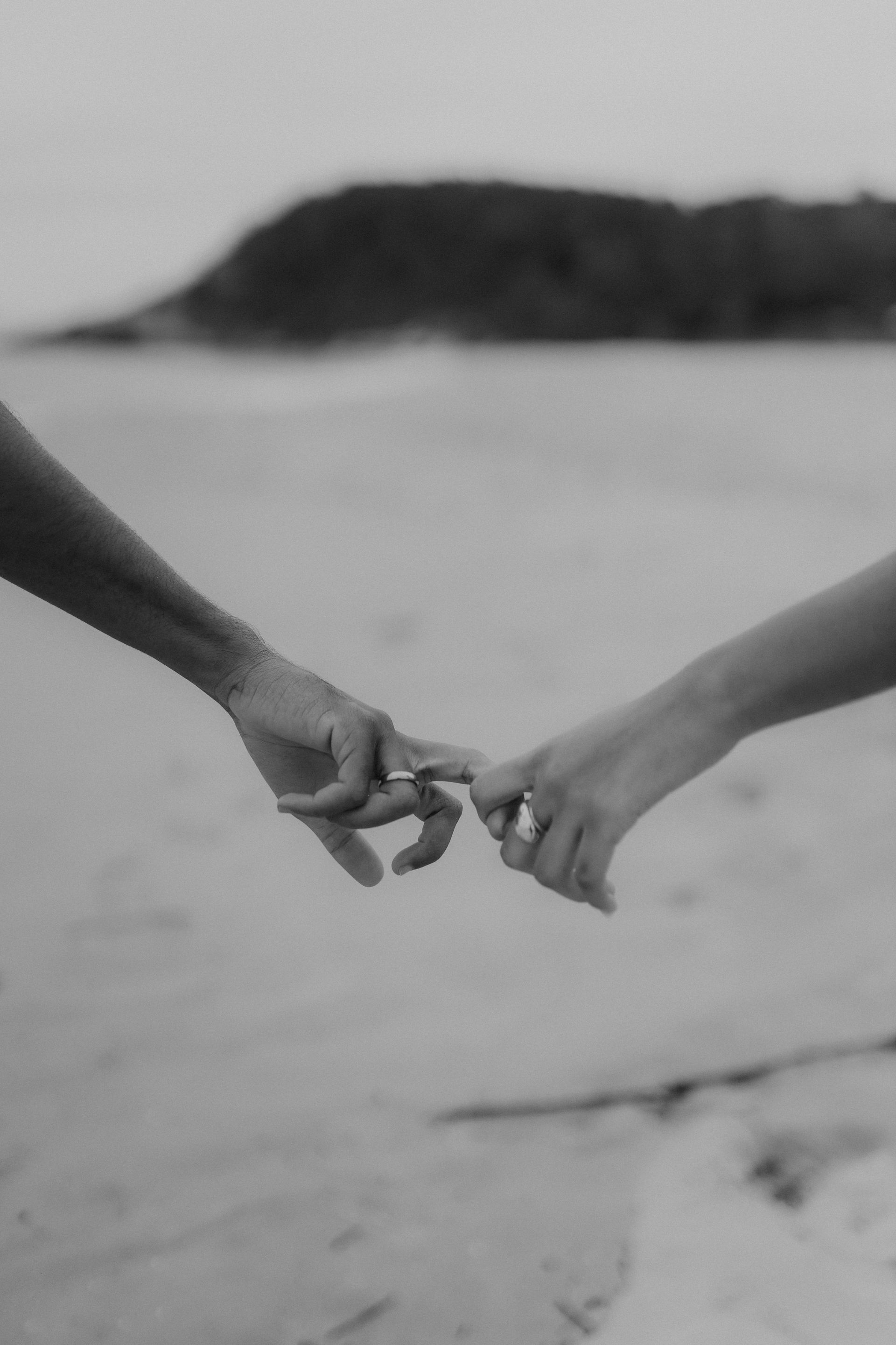 Two hands, fingers touching, pinky promise on beach, grayscale.
