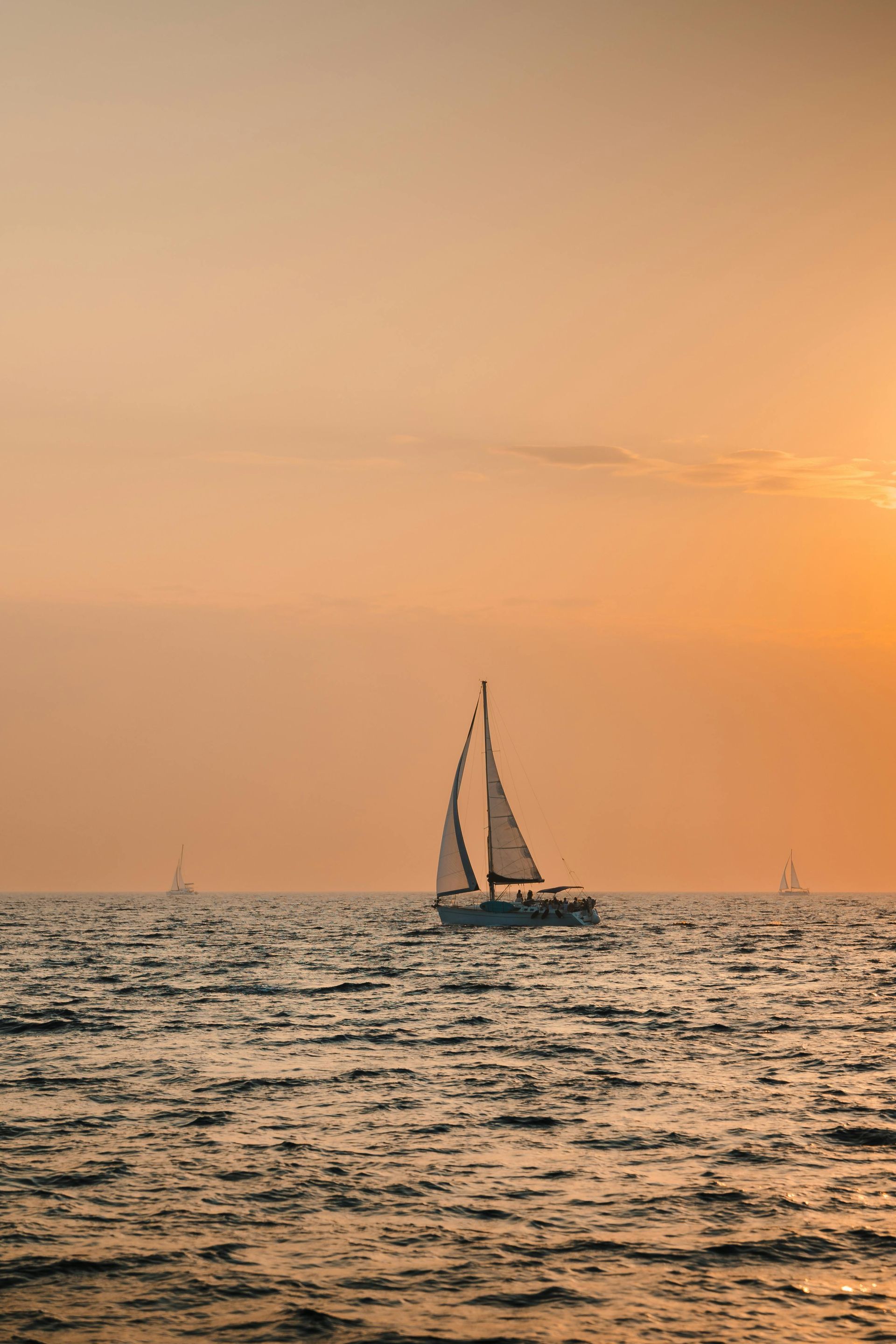 Sailboat on ocean at sunset, orange sky, small boats in the distance.