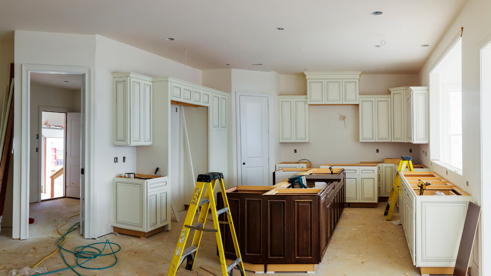 Kitchen renovation in progress: white cabinets installed, island with brown cabinetry, ladder presen