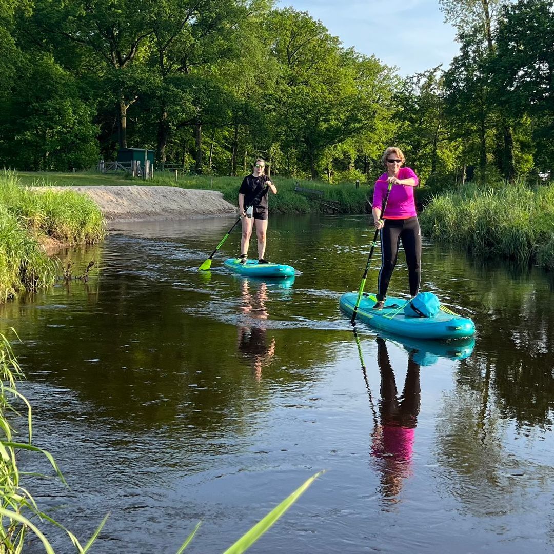 Twee mensen suppen op een rustige, smalle rivier, omzoomd door weelderige groene struiken en planten, onder een bewolkte hemel.