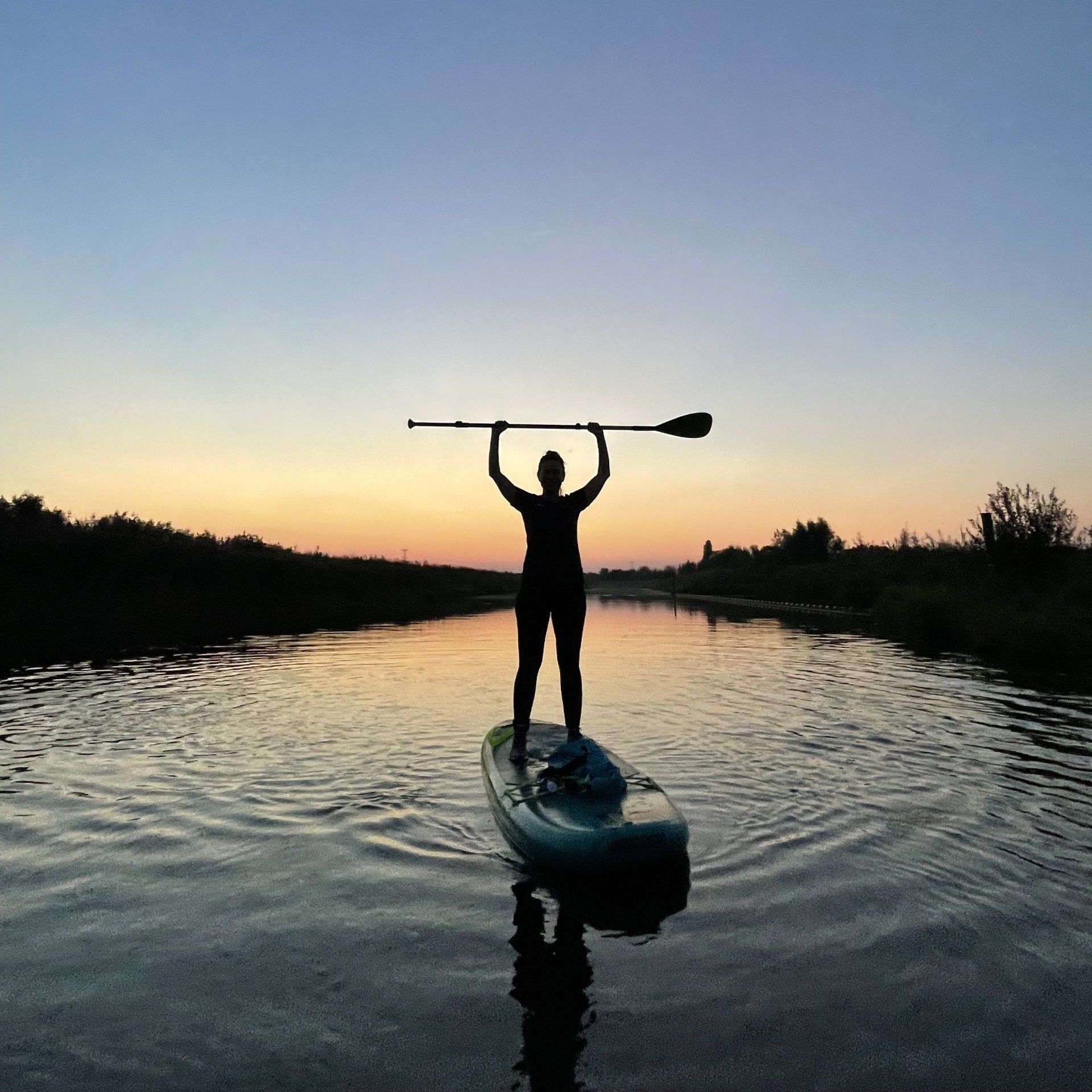 Een silhouet van een persoon die bij zonsondergang op een paddleboard in een rivier staat en een peddel horizontaal boven het hoofd houdt.