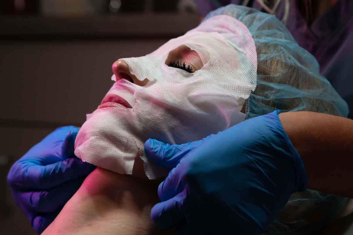 Person's face with a white sheet mask applied, held by gloved hands. Purple and blue lighting.