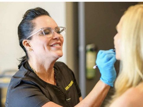 Woman in glasses smiles, examines patient's face with pen. Patient has blonde hair.