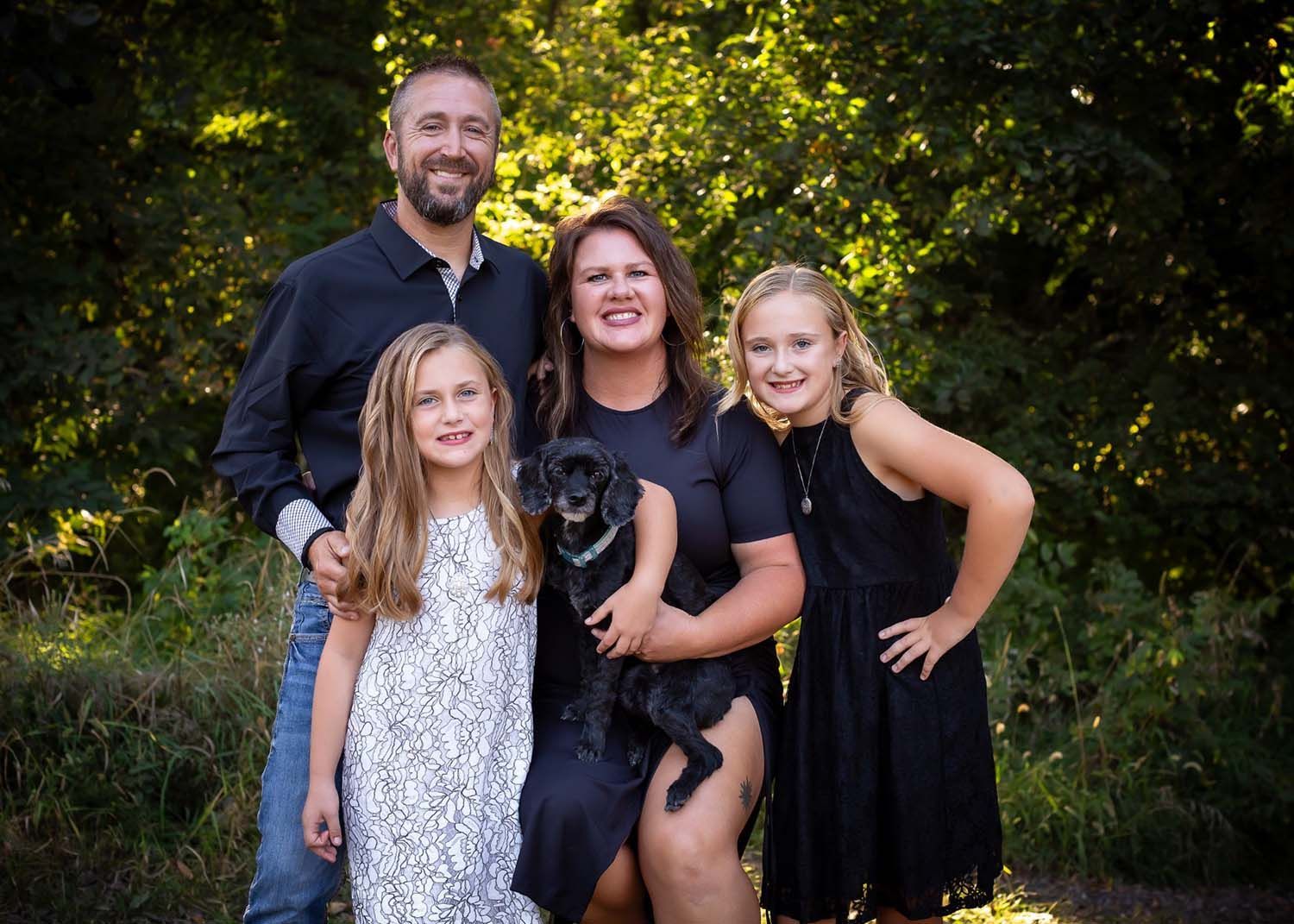 Family of four with a dog, posing for a photo. Man, woman, two girls, and a black dog outside with foliage.