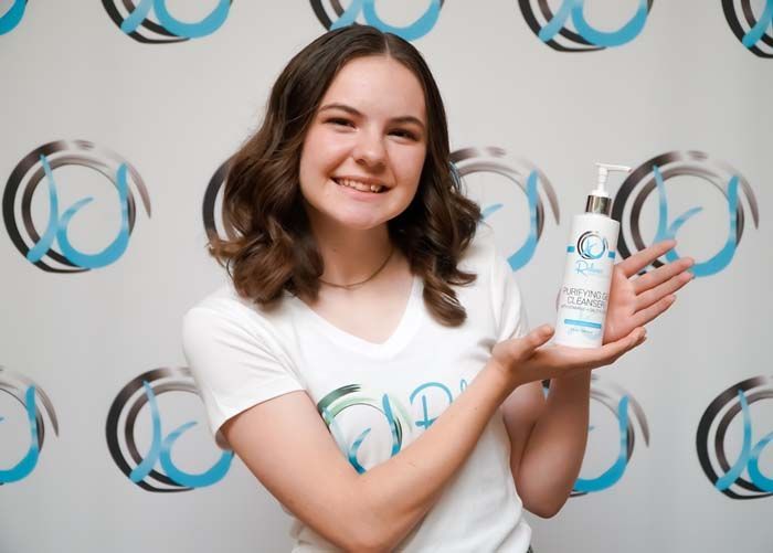 Woman holding a bottle of face cleanser, smiling at the camera. White shirt, logo background.