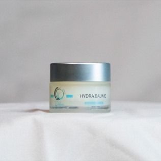 Small jar of Hydra Baume skincare product, silver lid, blue text, on white cloth.