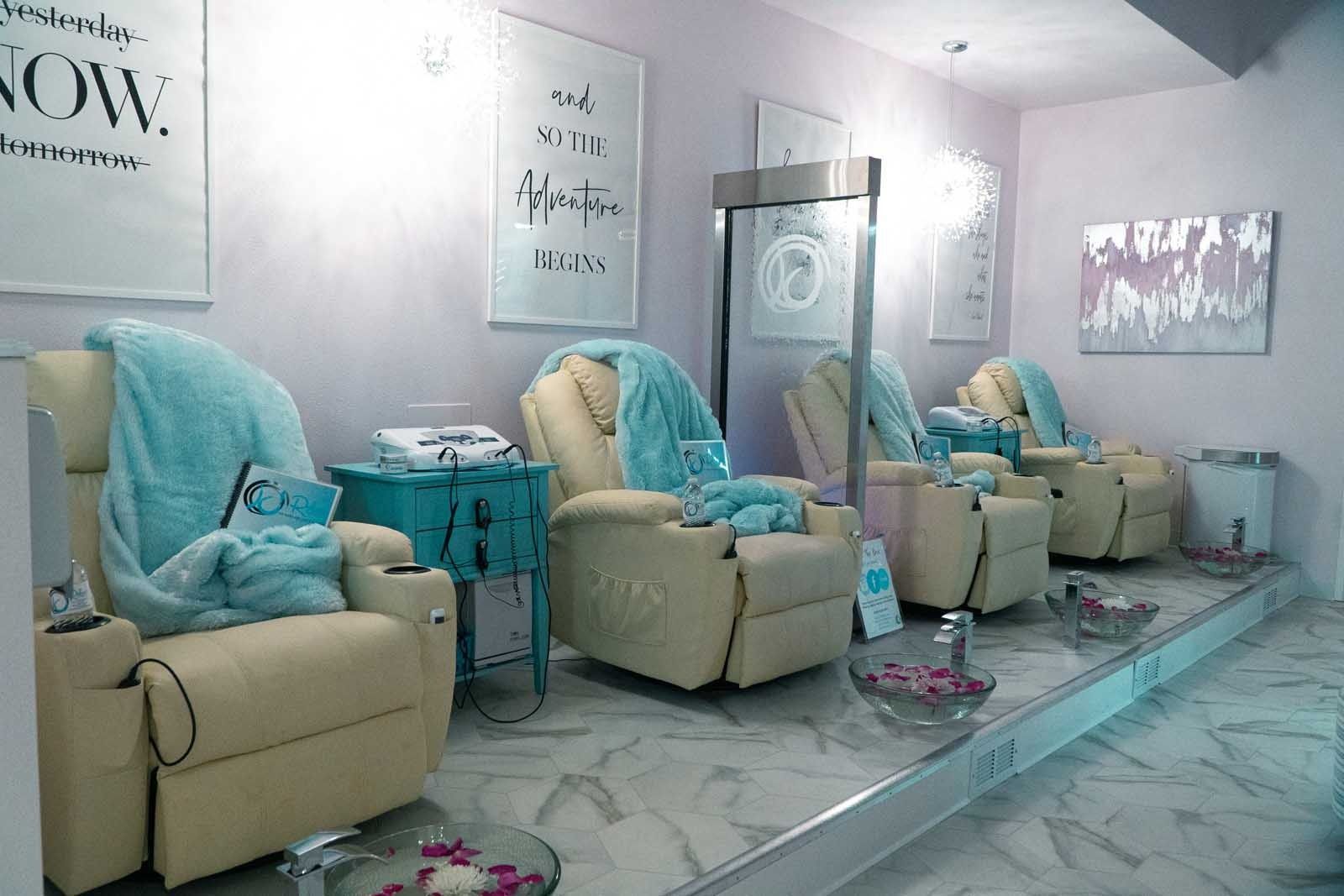 Interior of a nail salon with cream-colored reclining chairs, light blue blankets, and foot basins with rose petals.