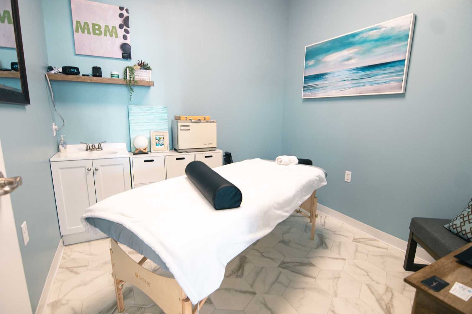 Massage therapy room with a massage table, artwork, and a sink, painted walls.