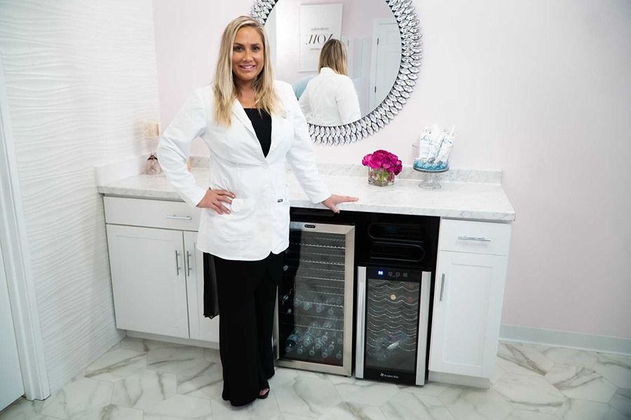 Woman in a lab coat, standing by a countertop with wine coolers, near a mirror in a light pink room.